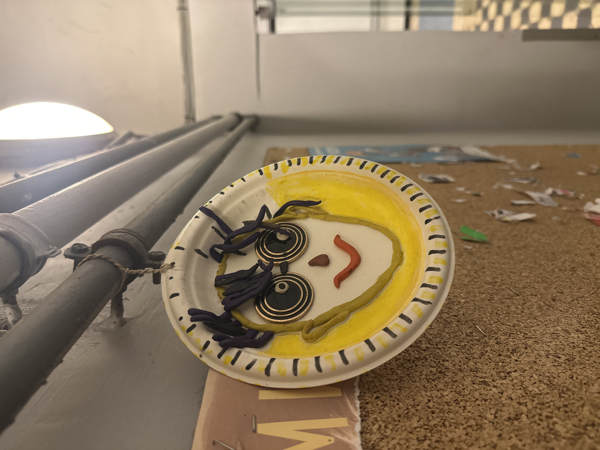 A paper plate decorated with clay is situated indoors. The plate displays a face with yellow "hair," round button eyes, and a small red clay mouth. The plate rests on a surface littered with debris and what looks to be part of a book cover. A pipe runs along the wall adjacent to the plate. There is no indication of the image being in Moscow, Russia.