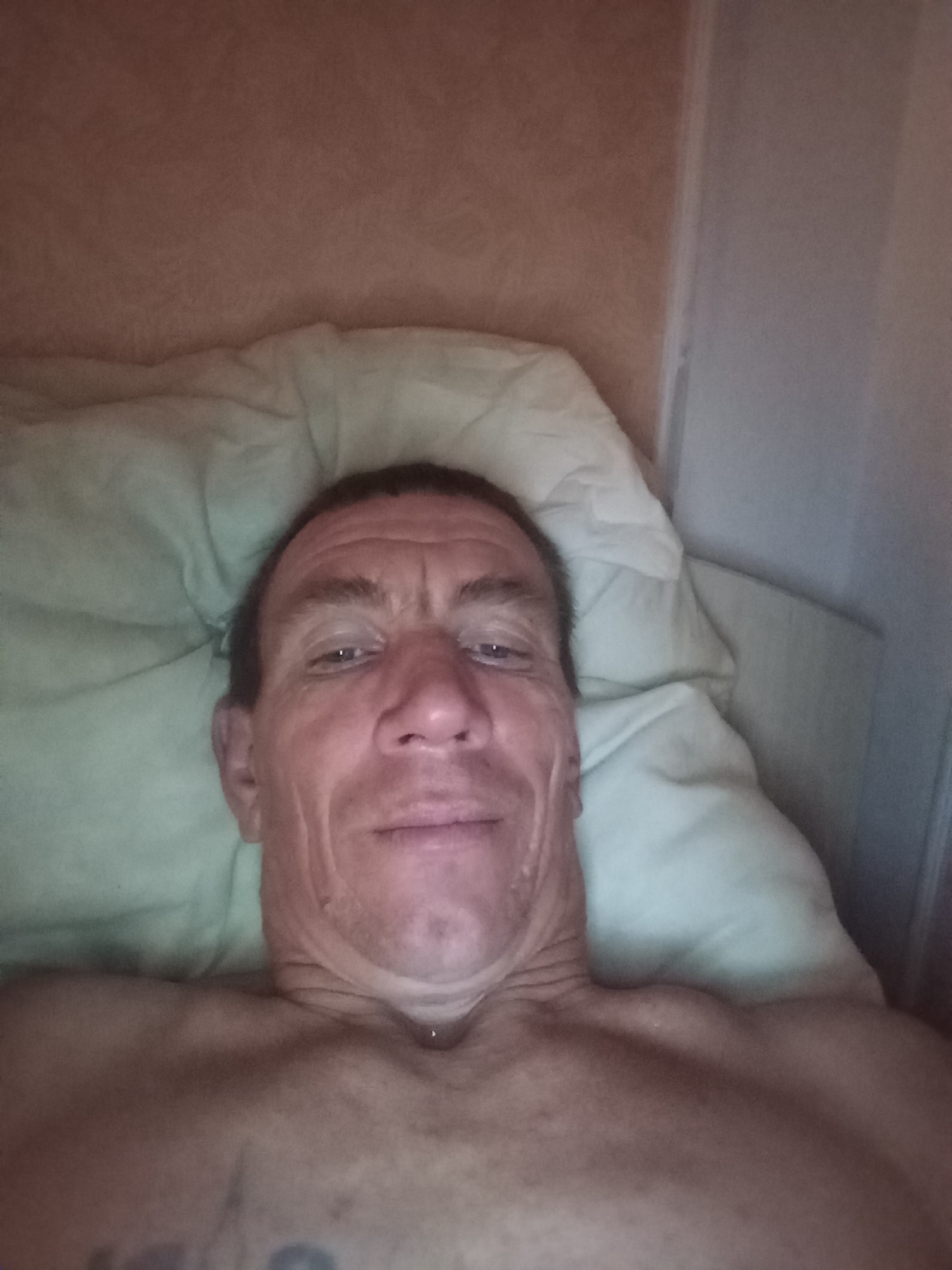 An elderly Caucasian male is lying on his back on a pillow. His head is tilted slightly back and to his left, with his face filling most of the frame. He has short, dark hair, fair skin with visible wrinkles, and is looking directly at the camera with his eyes open. The man is shirtless, revealing his chest and shoulders. The background consists of a light-colored wall with a textured pattern and a wooden headboard. The lighting is dim and appears to be artificial. There is no indication of the specific location within Nefteyugansk, Russia, as the image only shows an indoor personal setting.
