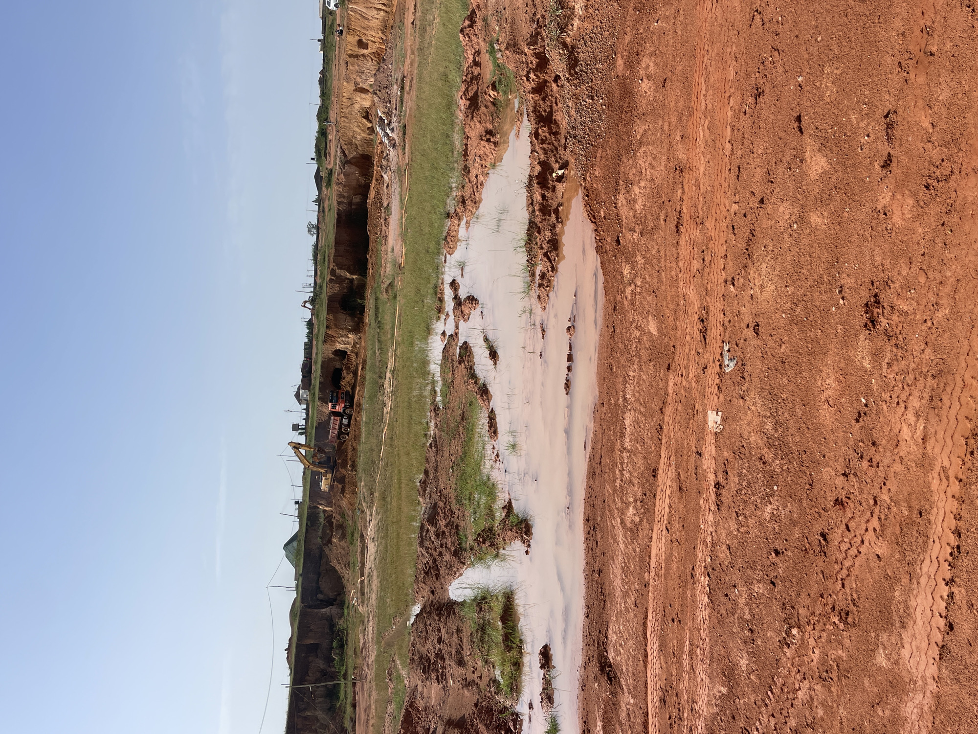 The image depicts a construction or excavation site in Jos, Nigeria. Predominantly red soil covers the ground, marked by tire tracks. A large pool of muddy water partially obscures the land, with tufts of grass and exposed earth interrupting the water's surface. On the left side of the image, a steep embankment reveals a cross-section of the soil, with construction equipment (possibly an excavator and a dump truck) visible along the upper edge of the excavation. In the background, buildings and vegetation are visible against a clear sky.