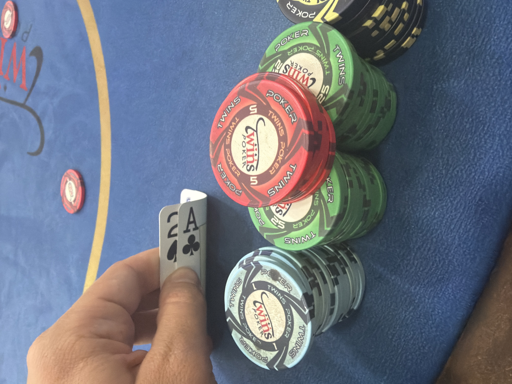 A person is holding two cards, the 2 of spades and the ace of clubs, next to stacks of poker chips of various colors. The poker chips have the text "TWINS POKER" on them. The setting appears to be a poker table with a blue felt surface. The image does not show the city of Larnaca, Cyprus.