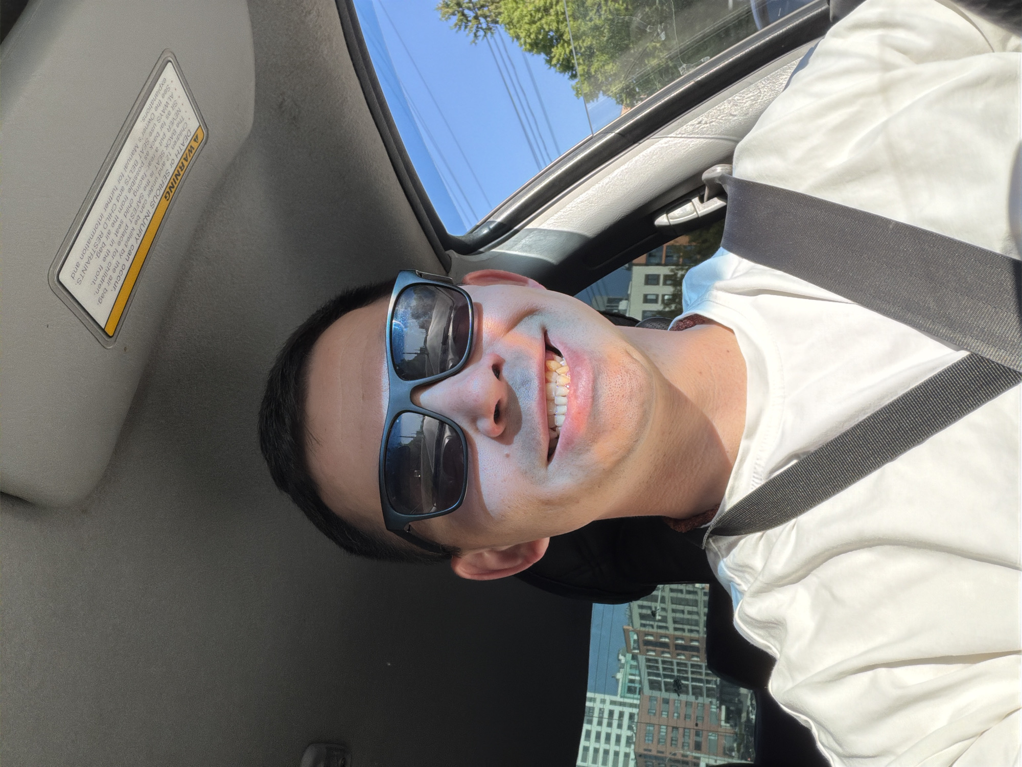 A selfie taken inside a car, likely in a city. The person, wearing sunglasses and a white shirt, is wearing a seatbelt. A sun visor with a warning label is visible in the upper left. Through the car window, buildings can be seen suggesting an urban environment. The exact location in Izhevsk, Russia, cannot be determined without additional information.