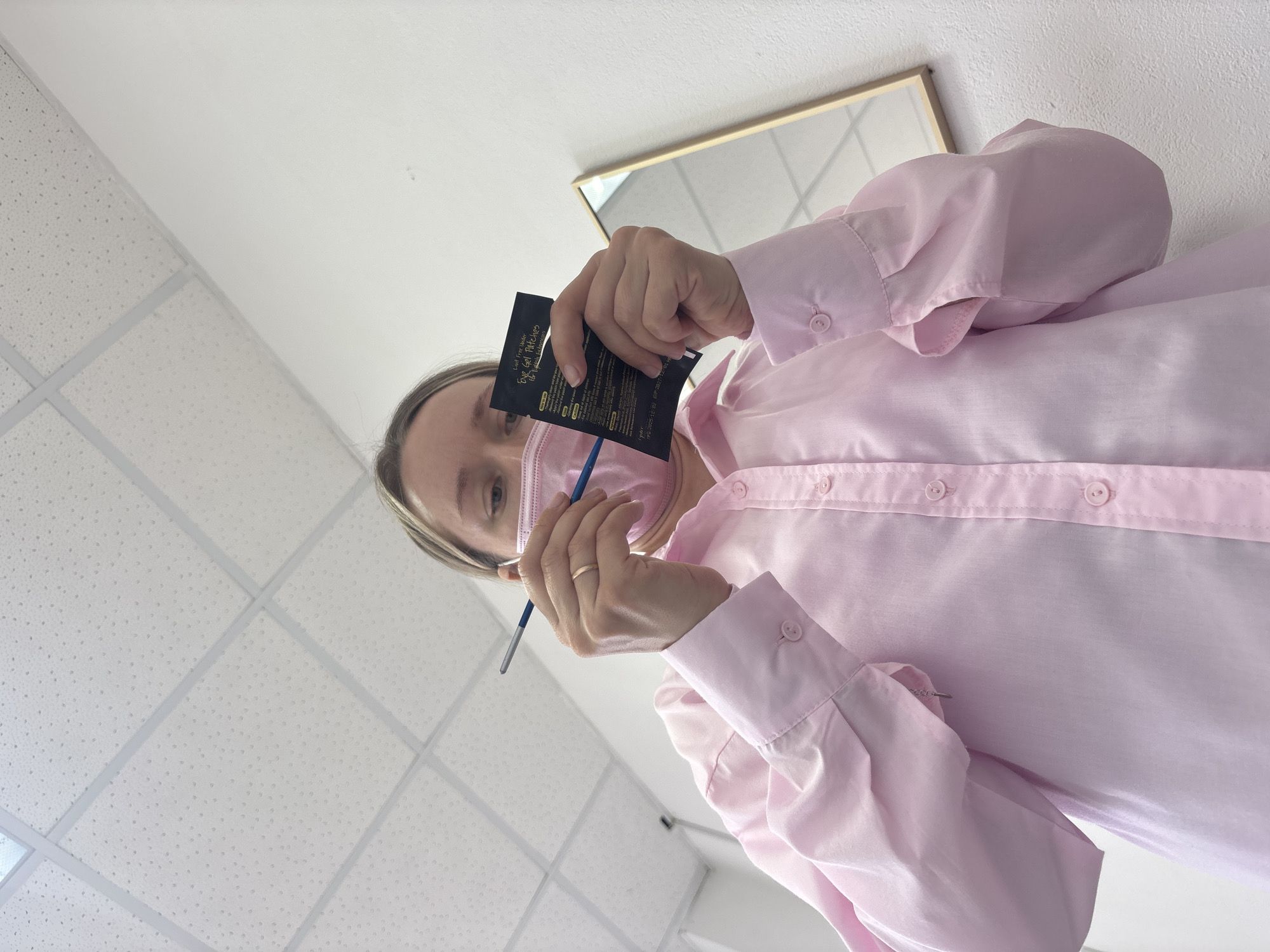 A woman wearing a pink button-up shirt and a pink face mask is holding a small black packet labeled "Lint Free Under Eye Gel Patches for Eyelash Extensions" and a blue-tipped applicator. The woman is looking down at the packet, and her hands are positioned to interact with it. The background consists of a white wall, a dropped ceiling with fluorescent lighting, and a reflection of the room in a framed mirror on the wall. The scene appears to be indoors, likely a personal care or beauty service location. The image is taken from a low-angle perspective. There is no discernible information in the image to indicate a location in Chaikovsky, Russia.
