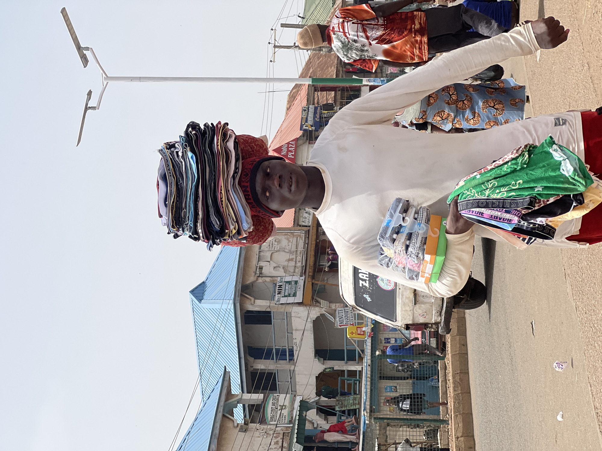 A man in Bukuru, Nigeria, balances a stack of folded clothing on his head and carries another stack of packaged goods. He is wearing a long-sleeved white shirt and a red hat. He is walking along a street in front of a multi-story building with businesses identified by signs like "Nim NIN New" and "NaNa PLAZA Collection". Street vendors can be seen. The street has a white light post.