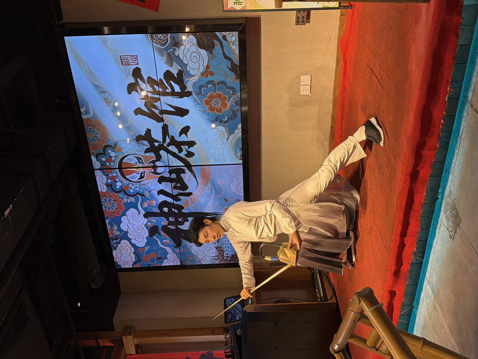 A male performer in a white traditional Chinese outfit, including a long-sleeved top and loose pants, is on a red-carpeted stage. He is in a lunging pose, holding a prop that resembles a watering can with a long spout. Behind him, a large screen displays a vibrant design featuring stylized clouds, flowers, and Chinese calligraphy, with an image of a Peking Opera mask. The performer's left leg is extended forward, and his right leg is bent. He is looking towards the audience, not visible in the frame. The stage is lit, and the background screen is the primary light source for the performer. Electrical outlets are visible on the wall behind the stage. A bamboo-like structure is in the foreground on the left. The scene appears to be a cultural performance in Yuzhong District, China.