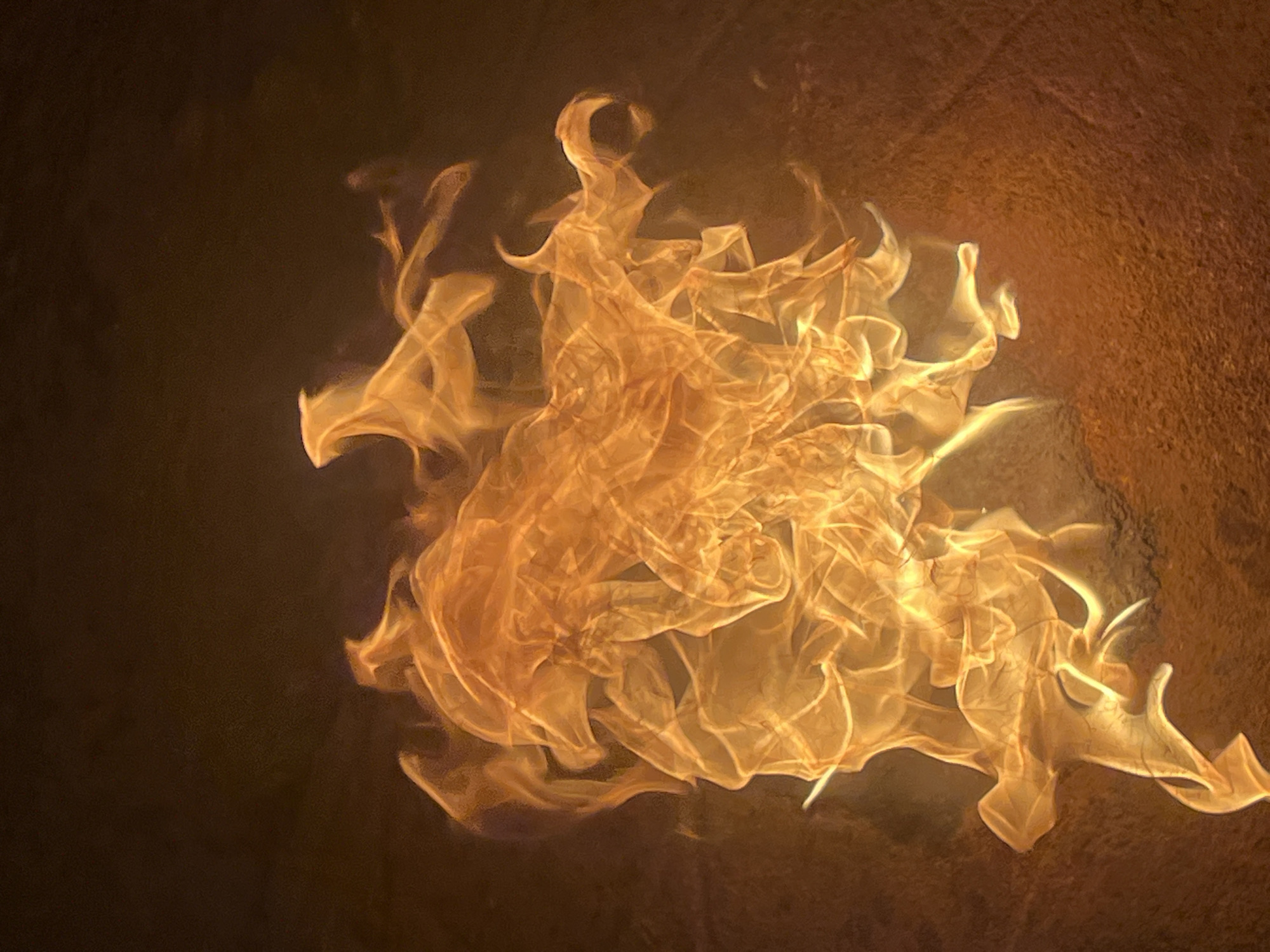 A close-up image features a cluster of flames, predominantly orange and yellow, against a dark background. The flames appear wispy and intricately shaped, filling a central area of the image. The background transitions from a darker shade on the left to a warmer, textured surface on the right. There are no people, objects, actions, or interactions visible. The location in Nkpor, Nigeria is not discernable from the visual data.