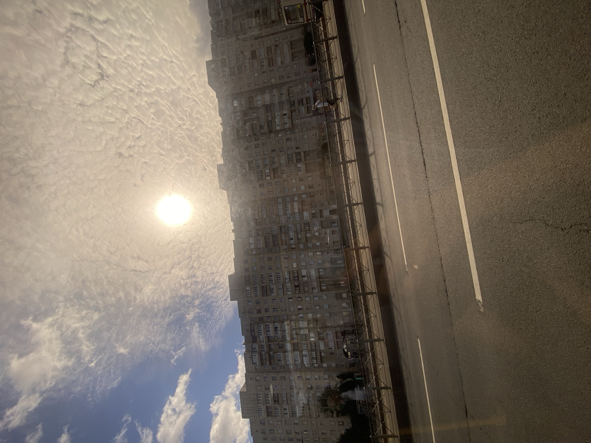 The image depicts a long, multi-story residential building characteristic of Soviet-era architecture in Saint Petersburg, Russia. The building is a uniform gray concrete structure with numerous balconies and windows. In the foreground, a road with white lane markings is visible, suggesting a street or highway. A pedestrian walkway with a metal railing separates the road from the building. The sky is partly cloudy with the sun prominently visible, casting a bright light and creating lens flares in the image. A few people are present on the walkway, and a bus is parked near the right side of the frame. The scene appears to be captured during the daytime.