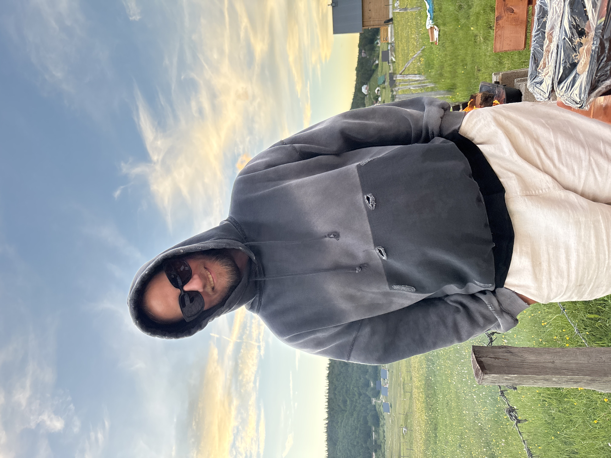 A man wearing sunglasses and a grey hoodie is standing outside in a grassy field with a barbed wire fence in the foreground. In the background, there are some wooden buildings, hills with trees, and a cloudy sky with hints of yellow and orange from what appears to be a sunset. The man has his hands in his pockets and is smiling at the camera. To the right of the man, there is a fire pit with a pot on it, and two containers covered in plastic wrap. This scene is located in Virak, Montenegro.
