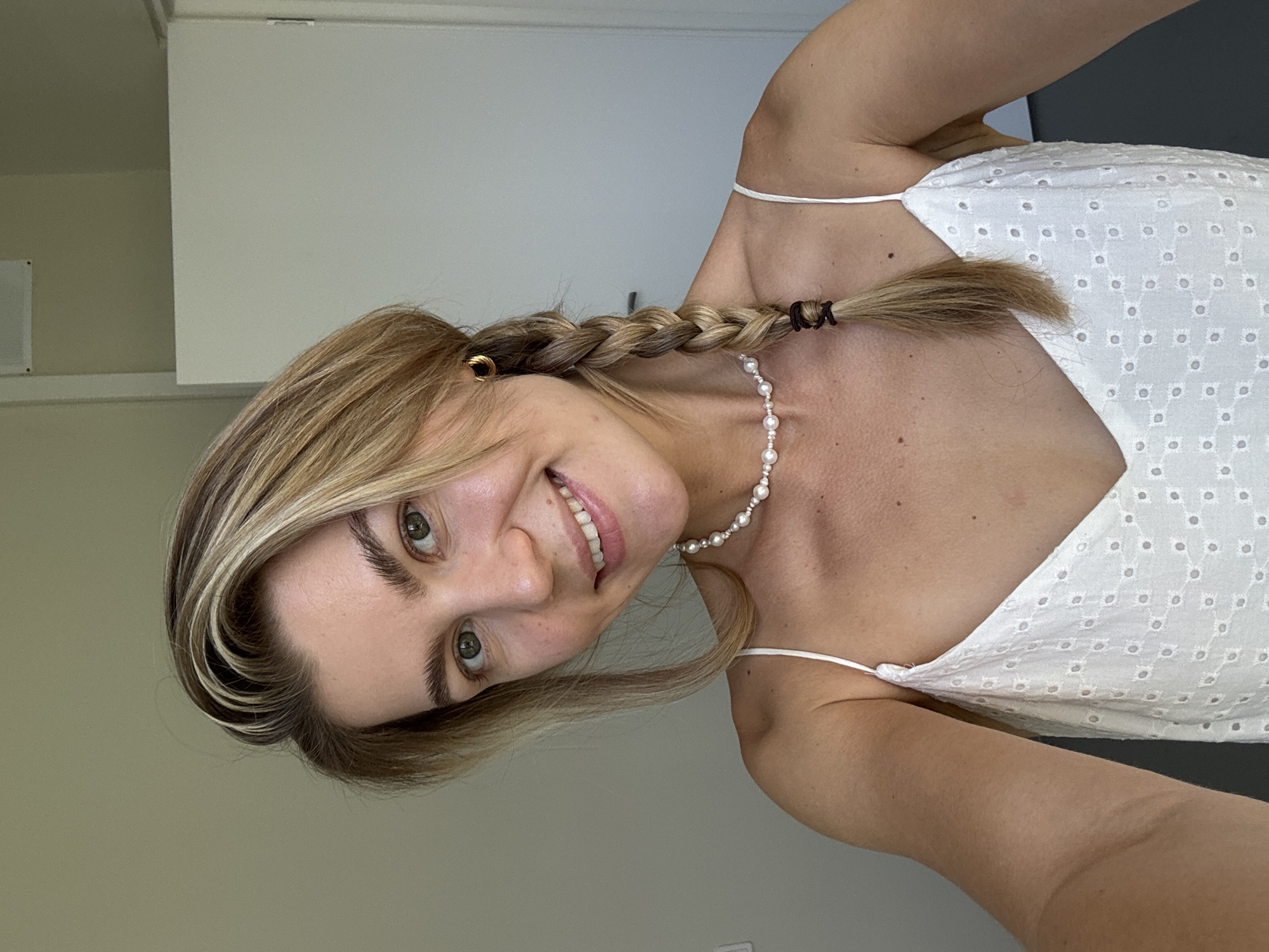 A young, blonde-haired woman with fair skin and green eyes is captured in a close-up, front-facing selfie. She is wearing a white, sleeveless dress with a delicate eyelet pattern and a pearl necklace. Her hair is styled in a braid, and she is wearing a small gold hoop earring. The woman is smiling directly at the camera, her head tilted slightly to the left. The background appears to be an indoor setting, with a plain wall and a door visible. The lighting suggests natural light coming from the right side of the frame. The overall impression is a casual, everyday portrait.