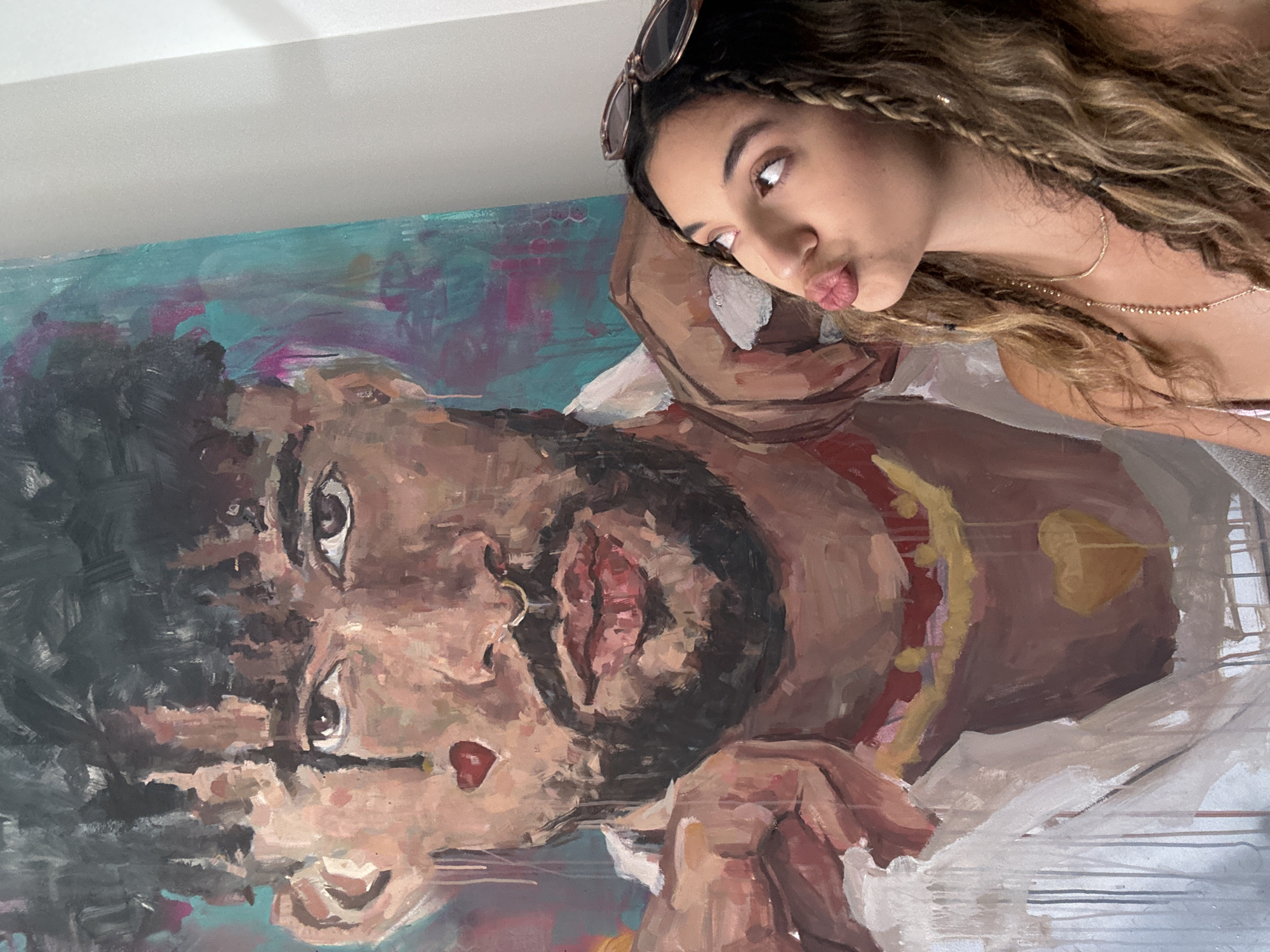A woman is posing in front of a large painting of a man. The painting is abstract and features bold brushstrokes and vibrant colors, with a teal and pink background. The man in the painting has dark, curly hair, a beard, and is wearing a white shirt with a red and yellow beaded necklace. He has a red heart shape painted on his cheek and a yellow heart shape on his chest. The woman, who has long, wavy brown hair with braids, is looking up and to the side with her lips pursed, as if about to kiss. She is wearing sunglasses on top of her head and a necklace. The painting appears to be quite large, taking up a significant portion of the background. The image is shot from a slightly low angle. The overall scene suggests a personal or artistic setting, possibly a home or studio. The question of location in San Juan, United States of America is not supported by any visual evidence in the image itself.
