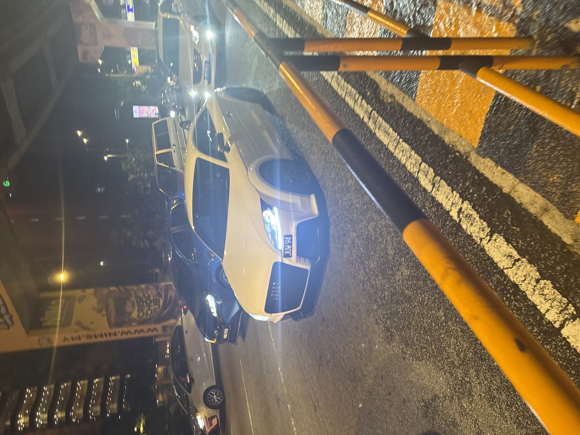 This nighttime image captures an urban street scene, likely in Kuala Lumpur, Malaysia. The central focus is a white Audi car, viewed from the front, with its bright LED headlights cutting through the darkness. The car's license plate, "JUW 94," is clearly visible on its front bumper.
To the immediate right of the Audi, a series of orange and black striped road barriers or poles are arranged, directing traffic or marking an area. The asphalt road surface, appearing somewhat reflective from the artificial lighting, features distinct white and yellow lane markings.
In the background, several other vehicles can be seen, some with illuminated lights, suggesting ongoing traffic or parked cars. On the far left, a large, dark elevated structure, possibly an overpass or bridge, frames the scene. Further back, various commercial signs and streetlights illuminate the environment, with one prominent billboard displaying the text "WWW.NIMS.MY" and "SOS DUIT". The overall setting is characteristic of a busy city at night, with no people visible and the weather appearing dry.
