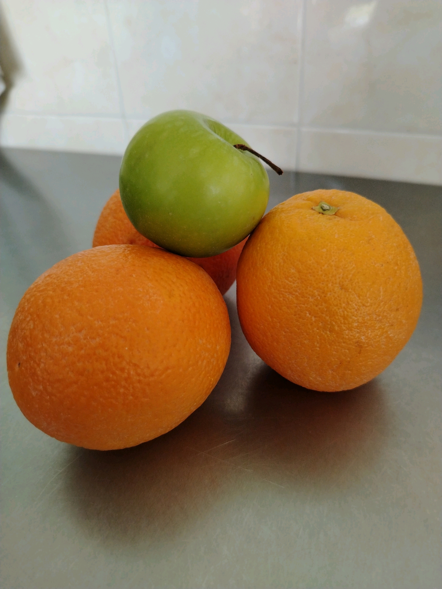 A close-up, eye-level shot shows a still life arrangement of three pieces of fruit on a metallic surface, likely a countertop. The fruits consist of one bright green apple and two round, orange-colored fruits, likely oranges. The apple rests atop two oranges, forming a small pyramid. The background is a light-colored tiled wall, with a subtle grid pattern. The lighting is even, casting soft shadows on the metallic surface. The image does not depict any people, actions, or interactions. It is a simple, well-lit composition of fruits. The location in Muravlenko, Russia, cannot be determined from the visual information provided.
