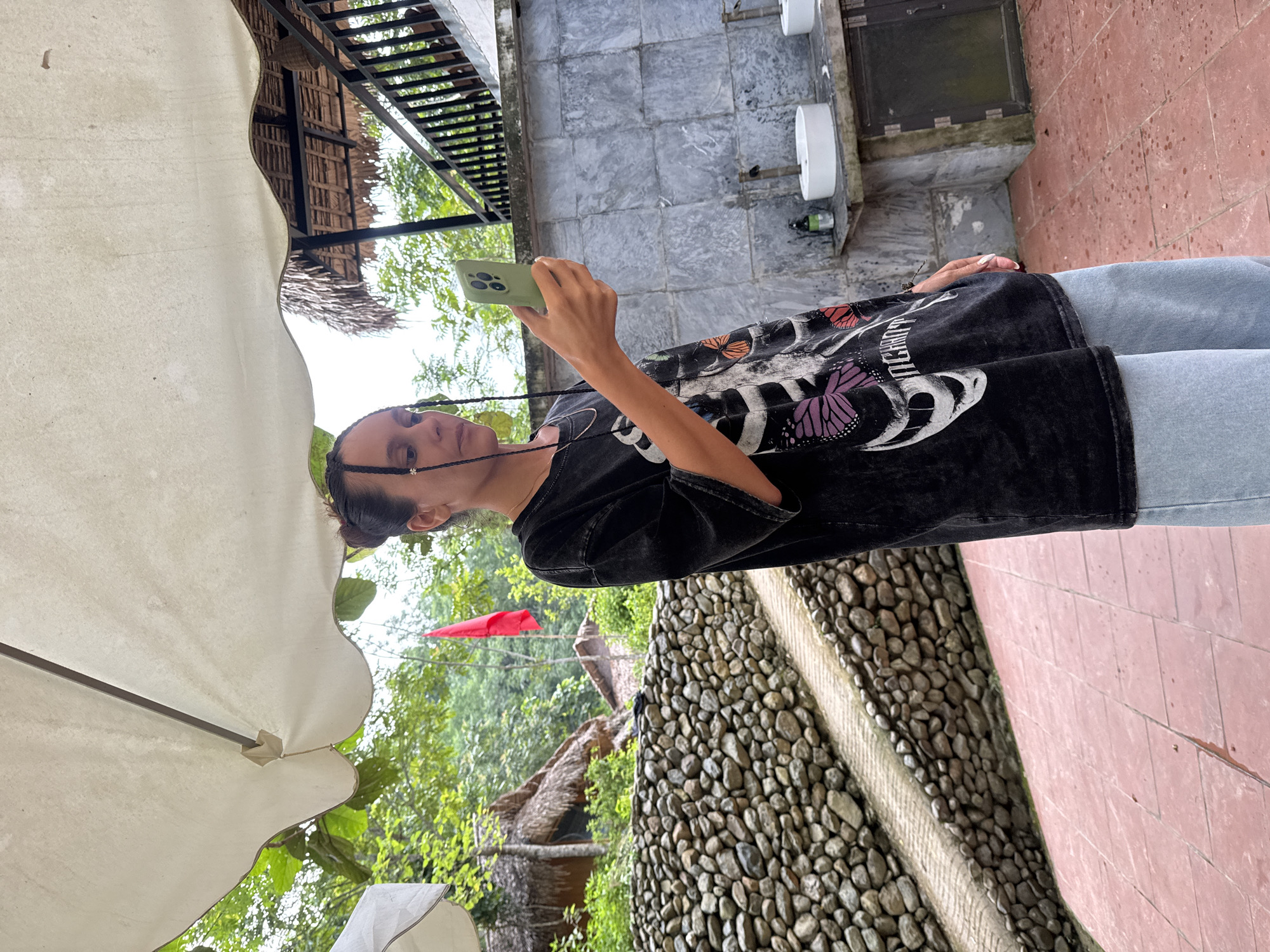 In Cổ Lũng Commune, Vietnam, a woman stands outdoors under a large white umbrella, holding a light green smartphone in her right hand and looking at its screen. She wears a dark t-shirt with a skeleton and butterfly graphic and light blue wide-leg pants. The background features lush green trees and vegetation. To the left, a textured stone wall with a paved pathway runs diagonally. To the right, a concrete structure with sinks and faucets is visible, along with dark railing and a thatched-roof building. A red flag is planted in the distance. The ground is paved with reddish-brown tiles.