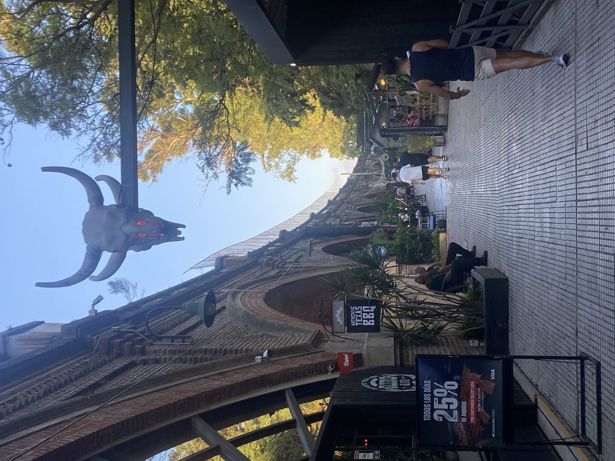 The image captures a street scene in Buenos Aires, Argentina, likely in the vicinity of the viaduct and associated structures. The view is angled upwards toward the viaduct, a brick structure with arched openings. The sky is visible above the viaduct. A large, gray, horned skull decoration is visible near the viaduct structure. Pedestrians are walking on the paved walkway in the foreground. There are signs for businesses, including one advertising "Authentic Texas BBQ" and another advertising "25% de ahorro" with Santander Select Visa, and the Rappi food delivery app. A man is jogging, and two people are sitting on a bench. Plants and trees are along the sidewalk.