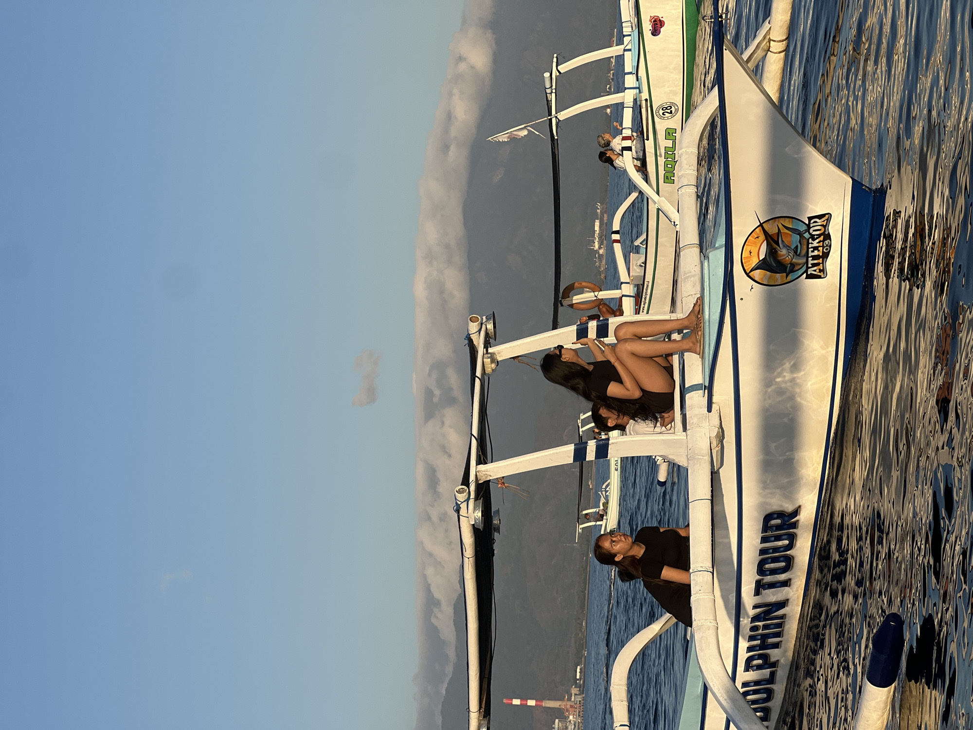 In the foreground, two boats are visible on the water. The boat on the left has the words "DOLPHIN TOUR" written on its side, along with a logo depicting a marlin. A woman in a black t-shirt and shorts sits on the left side of this boat, looking to the left. The boat on the right has the name "AQILA" and the number "28" on its side. Two women are seated on this boat, one in a black shirt and shorts, and another in a white shirt. In the background, there are mountains covered in clouds. A tall industrial chimney with red and white stripes is visible to the left of the mountains. The sky is clear and blue with a few wispy clouds. The water reflects the sky and the boats. The overall scene suggests a tour activity taking place at sea.