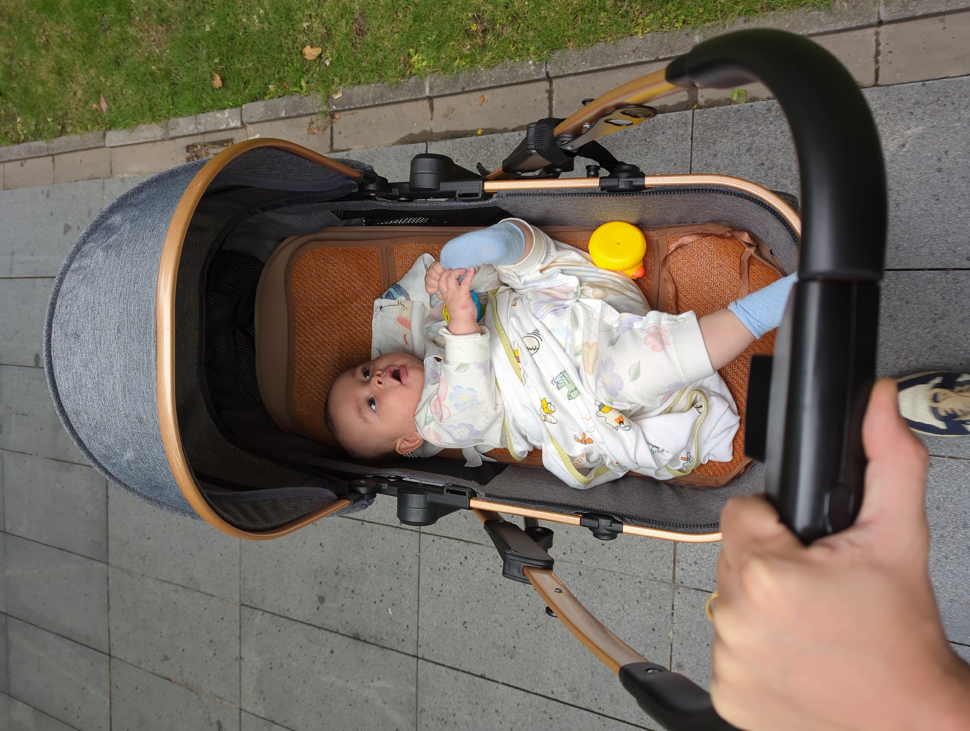 A top-down view shows a baby in a pram being pushed on a paved path. The baby is lying on its back, wearing a white patterned onesie and blue socks. A yellow toy duck is beside the baby. A hand holds the pram handle, and part of a shoe is visible. The path is bordered by grass and a low concrete wall, indicating an urban area. The scene does not inherently provide specific details about the location within Shunde District, China.