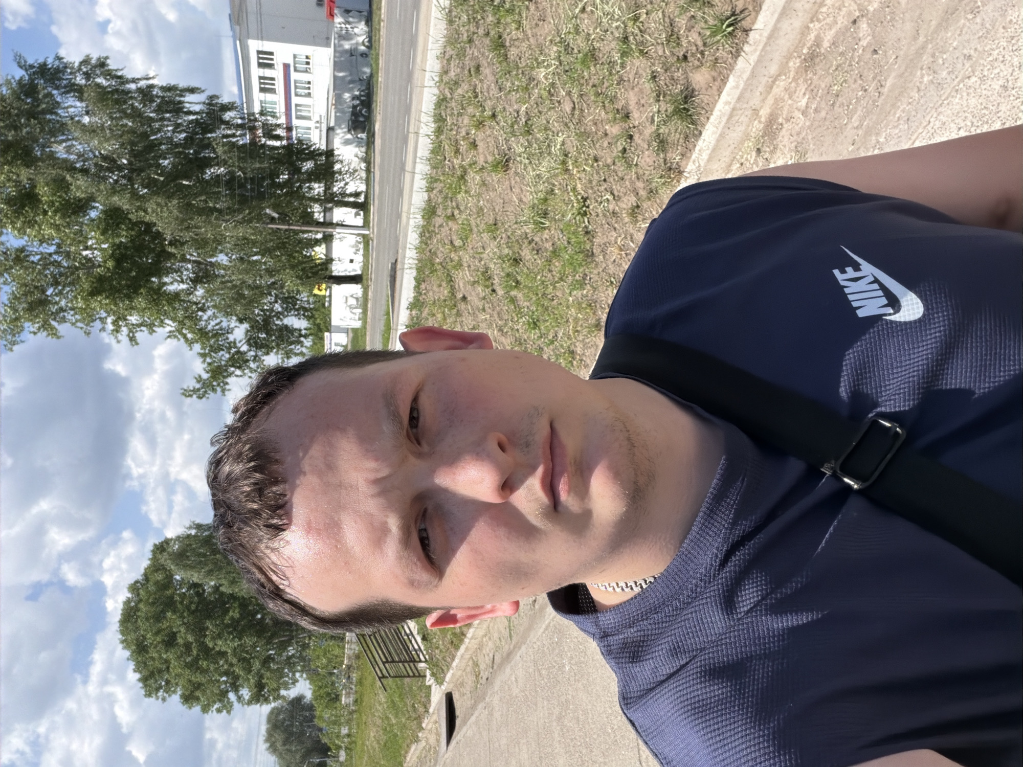 A selfie taken outdoors during daylight shows a fair-skinned young man wearing a dark blue t-shirt with a white Nike logo on the right shoulder. He has short, dark hair and a silver chain around his neck. A black strap, likely from a bag, crosses his torso. The background includes trees, grass, a building with multiple windows, and a cloudy sky. The location is likely a sidewalk or path in Chaikovsky, Russia, based on the surroundings.