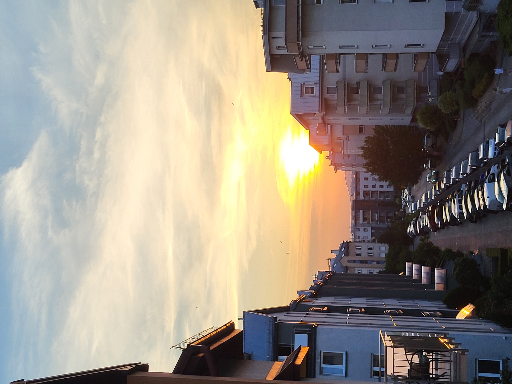 An aerial, eye-level, wide shot captures a street lined with apartment buildings and parked cars during a sunset. The sky is a gradient of pale blue at the top, transitioning to soft yellow and then a bright orange where the sun is setting. The sun itself is a bright, blown-out circle of light on the left side of the frame, casting a warm glow. Several wispy clouds are visible across the sky.
The apartment buildings on both sides of the street are modern in design, with balconies, windows, and flat or angled roofs. The building in the foreground on the left is partially visible, showing a balcony with outdoor furniture. The street is filled with parked cars, stretching into the distance. The streetlights are on, casting pools of light on the pavement. Two people are visible walking on the sidewalk in the lower right corner of the frame. The overall impression is a tranquil urban scene at dusk.