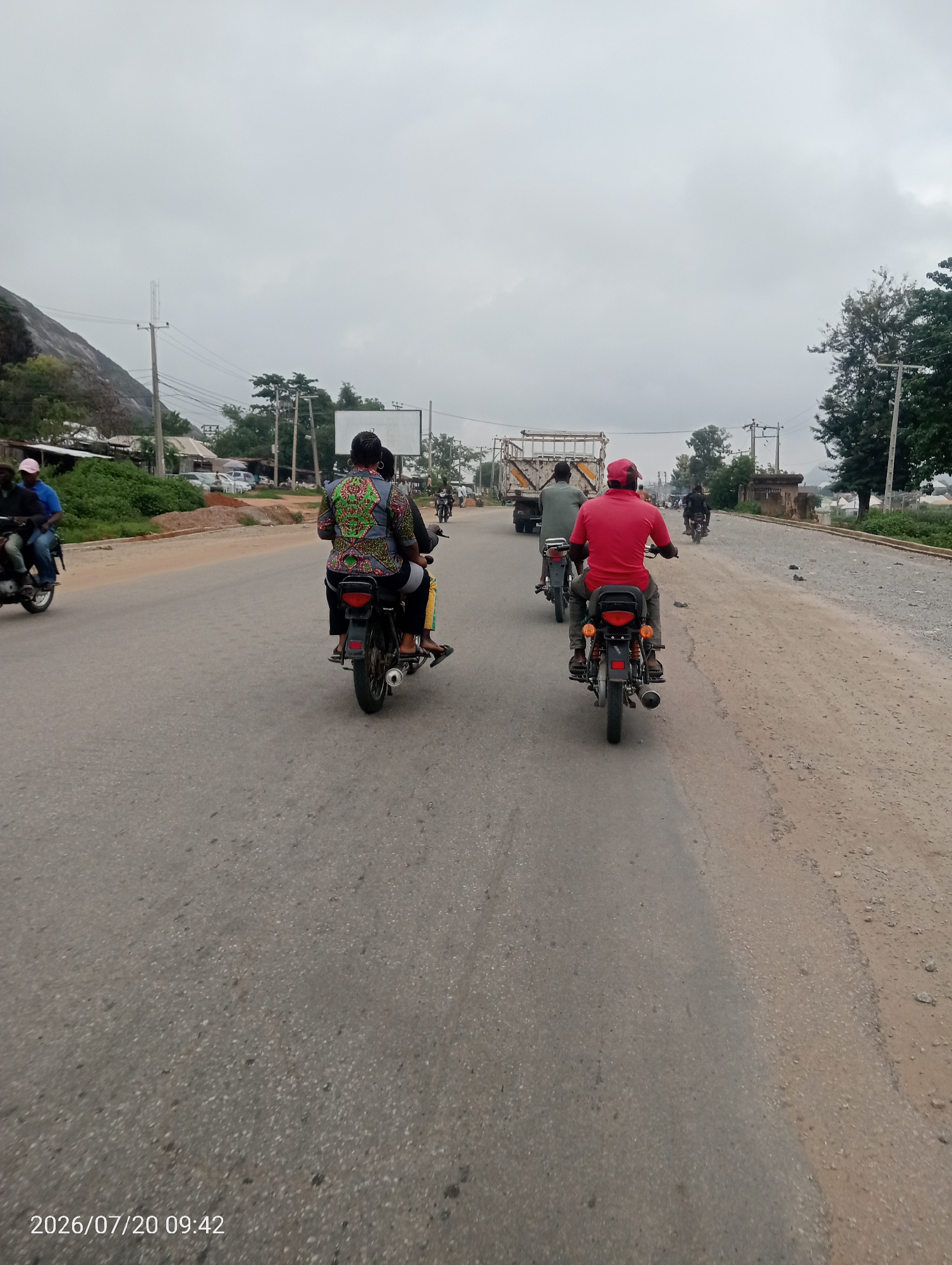 A perspective shot from a moving vehicle shows a road with several motorcycles and their riders. The road is paved and appears to be in decent condition. On the left side of the road, there are buildings and some vegetation, including trees and bushes. A rocky hill is visible in the background. On the right side of the road, there is a dirt shoulder with some gravel and sparse vegetation. The sky is overcast and cloudy. The image is dated July 20, 2026, at 09:42. This scene is consistent with the road conditions and environment found in Suleja, Nigeria.