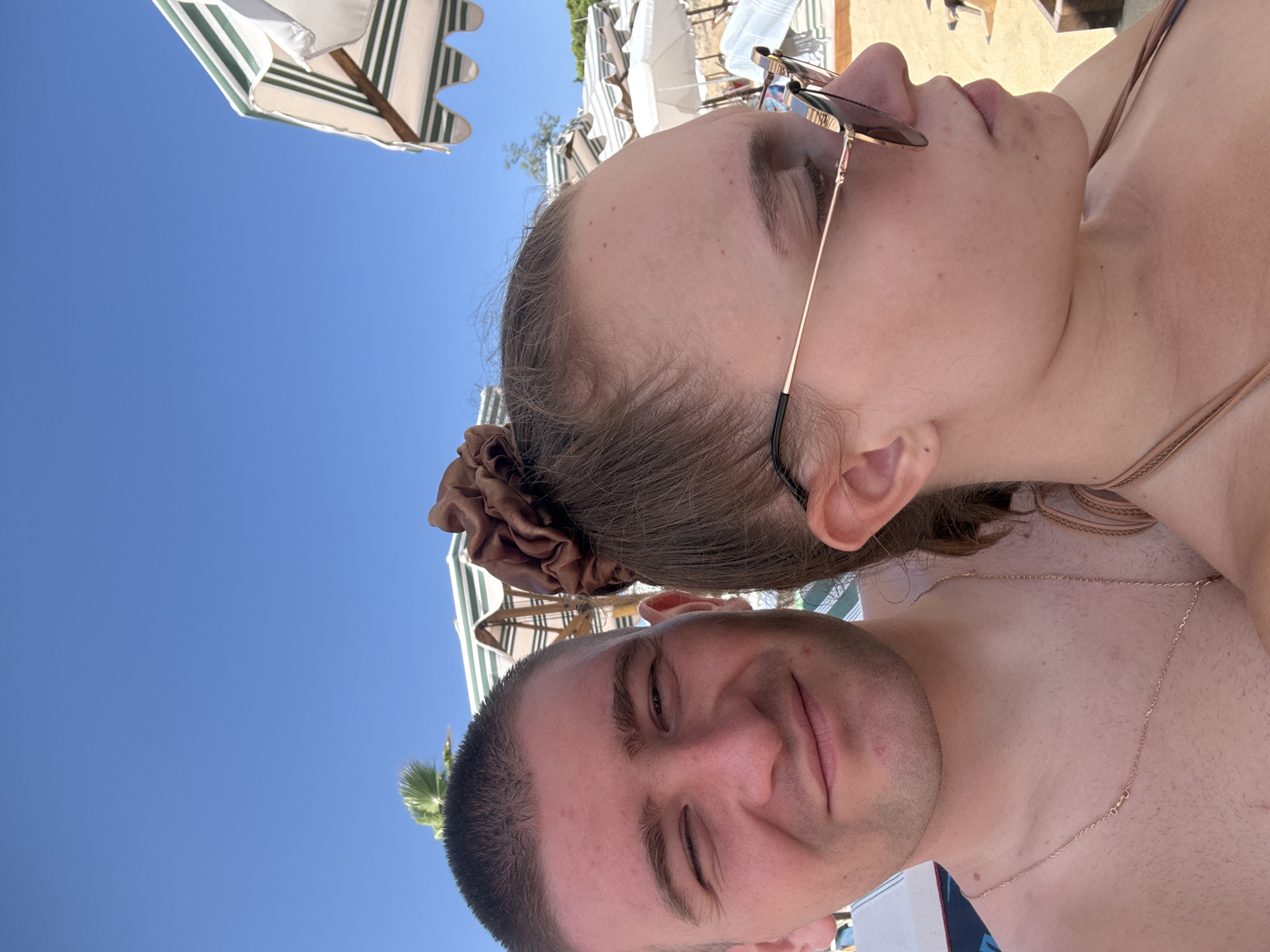 A close-up, wide-angle selfie shows a young man and a young woman at the beach under a clear blue sky. The man, on the left, has short hair and is winking. He is bare-chested and wearing a thin gold necklace. The woman is on the right, facing away from the camera and looking to the side. She has brown hair pulled back in a scrunchie and is wearing sunglasses and a bikini top. In the background, beach umbrellas with green and white stripes are visible, along with palm trees. The overall scene suggests a sunny day at a beach resort.