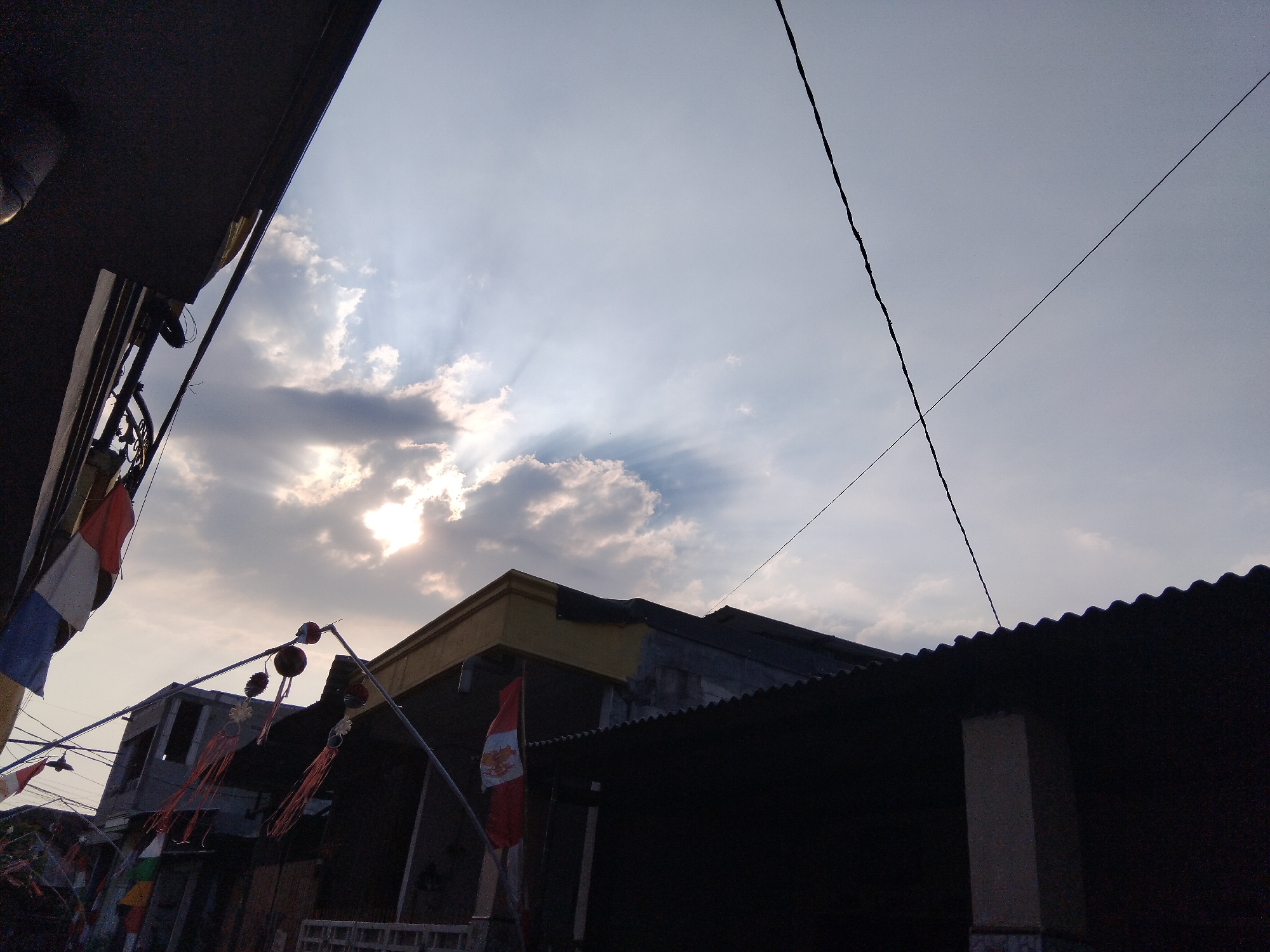 The image captures a view of buildings and the sky in Kepuh Kiriman, Indonesia. In the foreground, on the left side, a dark overhang juts into the frame. Below this, a series of flags, one with red, white, and blue stripes and another with red and white, are visible, hanging from wires. Further into the scene, several buildings are depicted. One building has a yellow trim at the top and appears to be under construction or in disrepair, with exposed brickwork. Another building to the right has a dark, corrugated metal roof. Various wires crisscross the sky, and decorative elements, possibly lanterns or tassels, are suspended between poles in front of the buildings. The sky is filled with clouds, and the sun is breaking through, creating visible sun rays. The overall lighting suggests it is either sunrise or sunset.