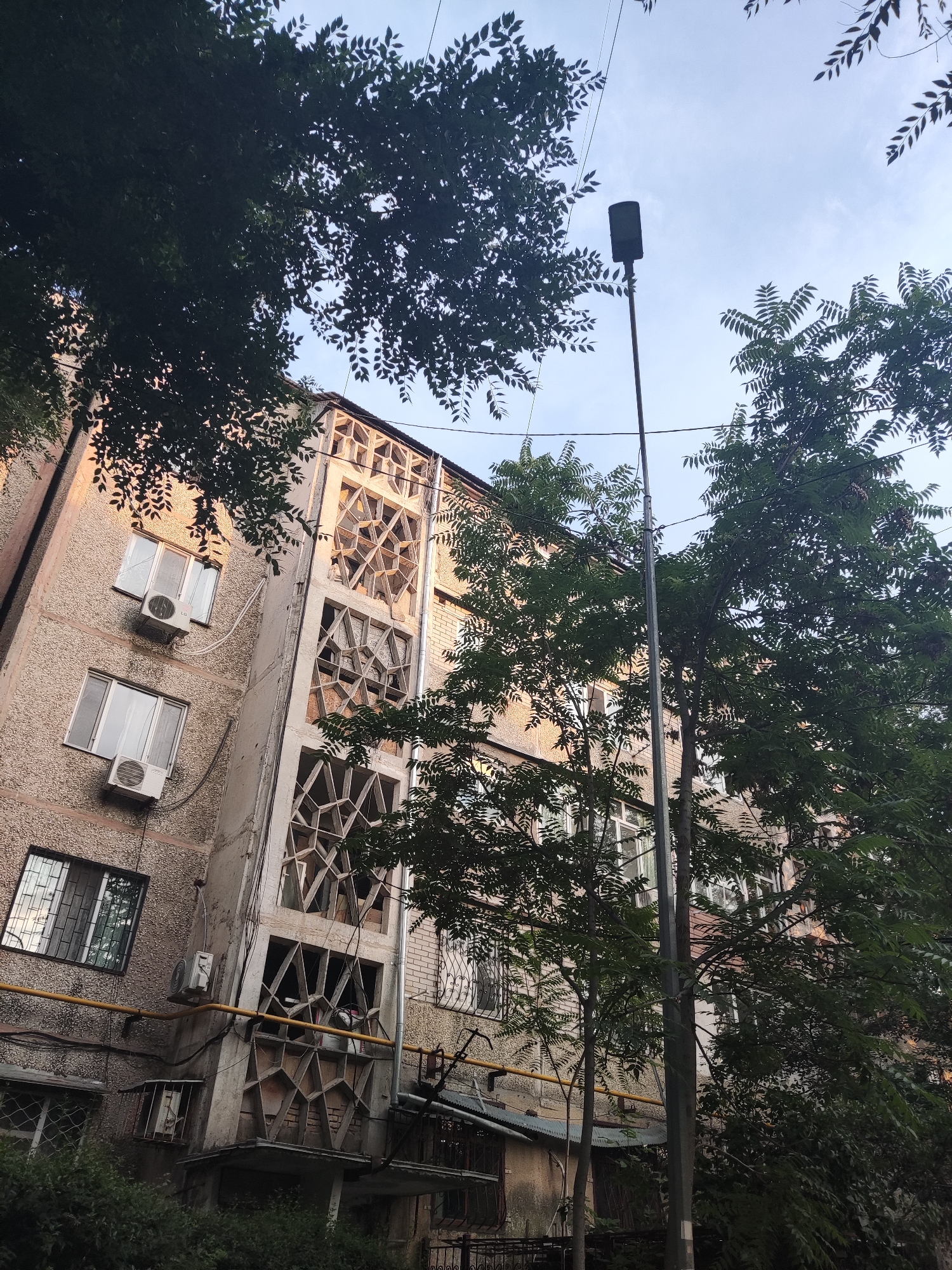 This media file captures a street-level view of an apartment building in Shymkent, Kazakhstan. The building's facade is textured and appears to be made of concrete, with various windows and air conditioning units visible. A distinctive architectural feature is a decorative concrete structure with geometric, angular openings, possibly a balcony or an accent wall, on the right side of the building. A yellow pipe runs horizontally across the lower portion of the building.
Several trees with dense green foliage partially obscure the building and the sky, suggesting an outdoor setting during daylight. The sky is visible through the leaves and is a pale blue, indicating it could be daytime with some cloud cover or early morning/late afternoon light. A street lamp stands tall near the center-right of the frame. The overall impression is that of an urban residential environment. There is no visible text in the image. The mood is neutral, depicting a common urban scene.