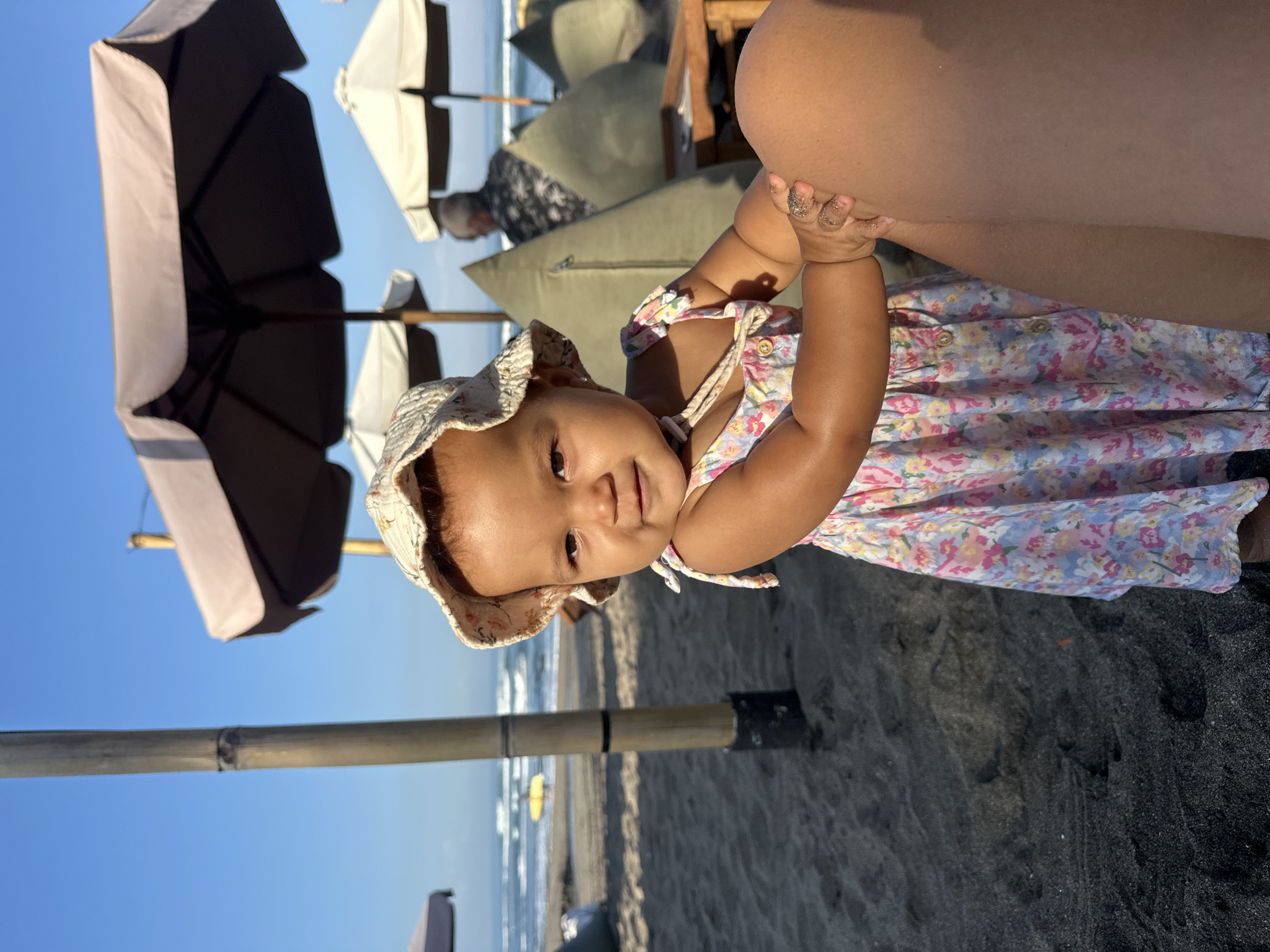 A young child wearing a floral dress and a sun hat smiles at the camera on a dark sand beach in North Kuta, Indonesia. The child's right hand rests on an adult's leg. In the background, beach umbrellas and lounge cushions are visible, along with the ocean and sky.