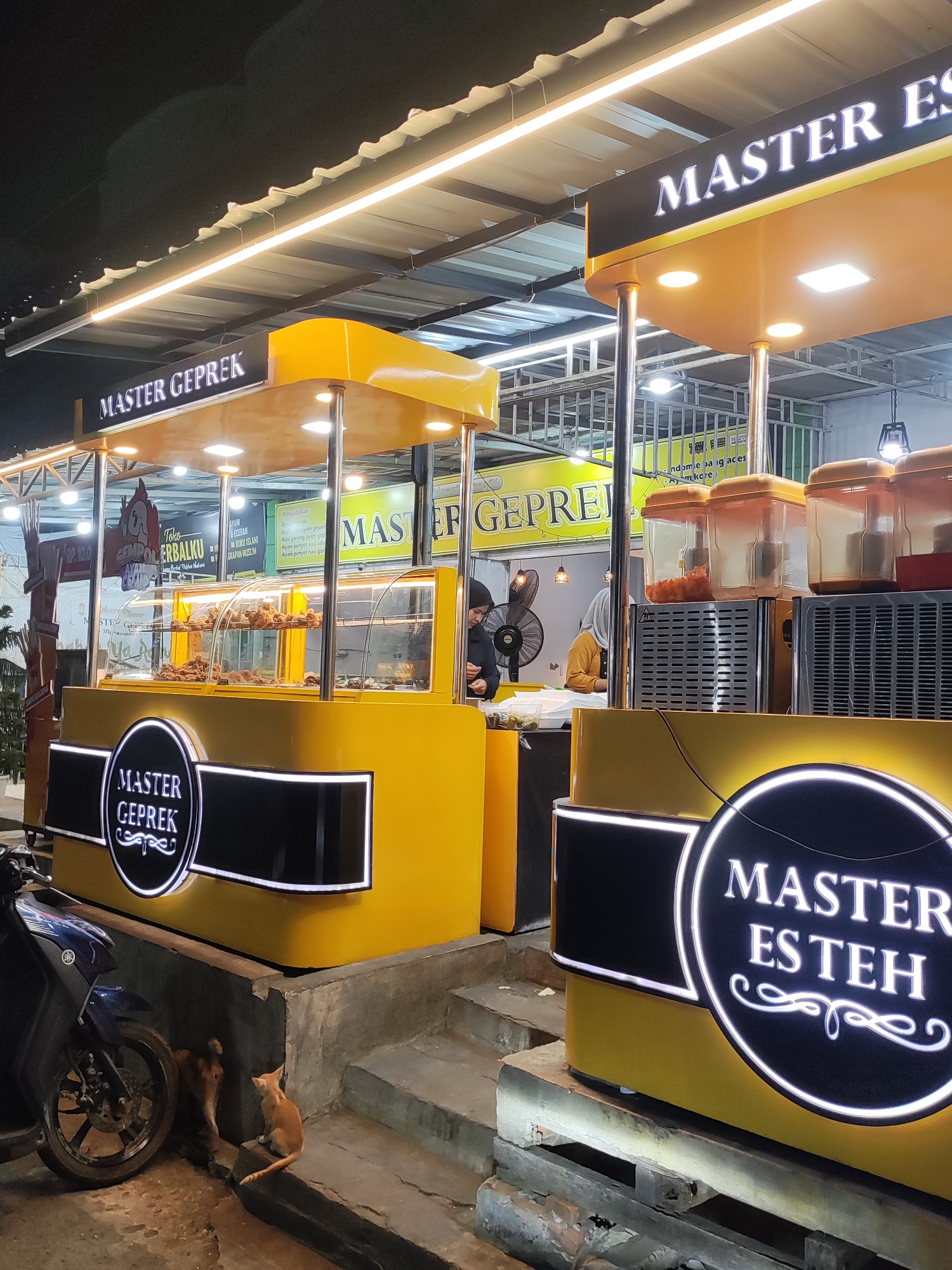 A vibrant nighttime street scene in Lhokseumawe, Indonesia, features two brightly lit yellow and black food stalls operating under an awning. The primary stall on the left, prominently labeled "MASTER GEPREK," has a glass display case showcasing numerous pieces of fried chicken. A person in dark clothing, possibly a vendor, is visible working behind this counter. Above this stall, another sign for "Toko EMPOL ERBALKU" advertises "ECERAN BUKU ISLAMI" (retail Islamic books) and "PERLENGKAPAN MUSLIM" (Muslim supplies), alongside "AYAM" (chicken) and "RP. 10.0" (likely Rp. 10,000).
To the right, a second, partially visible stall is branded "MASTER ES TEH" (Master Iced Tea) with large, glowing white letters. This stall has several clear beverage dispensers on its top counter. Another vendor, wearing a yellow top and hijab, is visible behind this section. Bright strip lights illuminate the entire setup, contrasting with the dark night sky.
In the foreground, below the "Master Geprek" stall and beside a blue motorcycle, two cats are present: one orange cat is sitting and looking, while a darker cat is partially obscured. Other signs in the background, including one that reads "MASTER GEPREK" and lists menu items like "Ayam penyet nasi uduk" (smashed chicken with coconut rice) and "Ayam penyet nasi goreng" (smashed chicken with fried rice), further indicate the culinary offerings. The overall scene suggests a lively and casual outdoor dining experience.