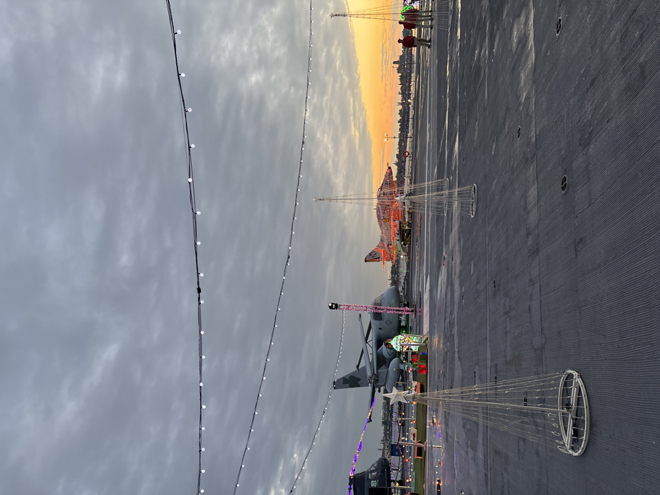 The image presents a festive scene on the flight deck of an aircraft carrier, likely the USS Midway Museum in San Diego, during the transition from dusk to evening. The sky overhead is heavily overcast with grey clouds, but a vibrant orange and yellow sunset is visible on the horizon, casting a warm glow over a distant city skyline and a body of water.
The main subjects are the Christmas decorations and the aircraft on the deck. Strings of white lights are strung across the top of the frame, creating an overhead canopy. Several large, conical light sculptures, made of white frames and embedded lights, are scattered across the deck, one in the foreground topped with a white star.
Two aircraft are prominent: a fighter jet, fully covered in bright orange and red Christmas lights, is positioned towards the rear of the deck. Closer to the foreground and to the left, a large helicopter, possibly a CH-46 Sea Knight, is also decorated, with a festive, illuminated gift box-like structure placed near its base.
In the far background, two silhouetted individuals are visible on the deck, observing the sunset. The scene depicts a holiday event, transforming the military vessel into a public attraction with seasonal illuminations. The time of day is clearly sunset or early twilight, creating a dramatic and atmospheric setting. The weather appears cloudy, with a distinct break in the clouds at the horizon for the sunset. No visible text is present in the image.