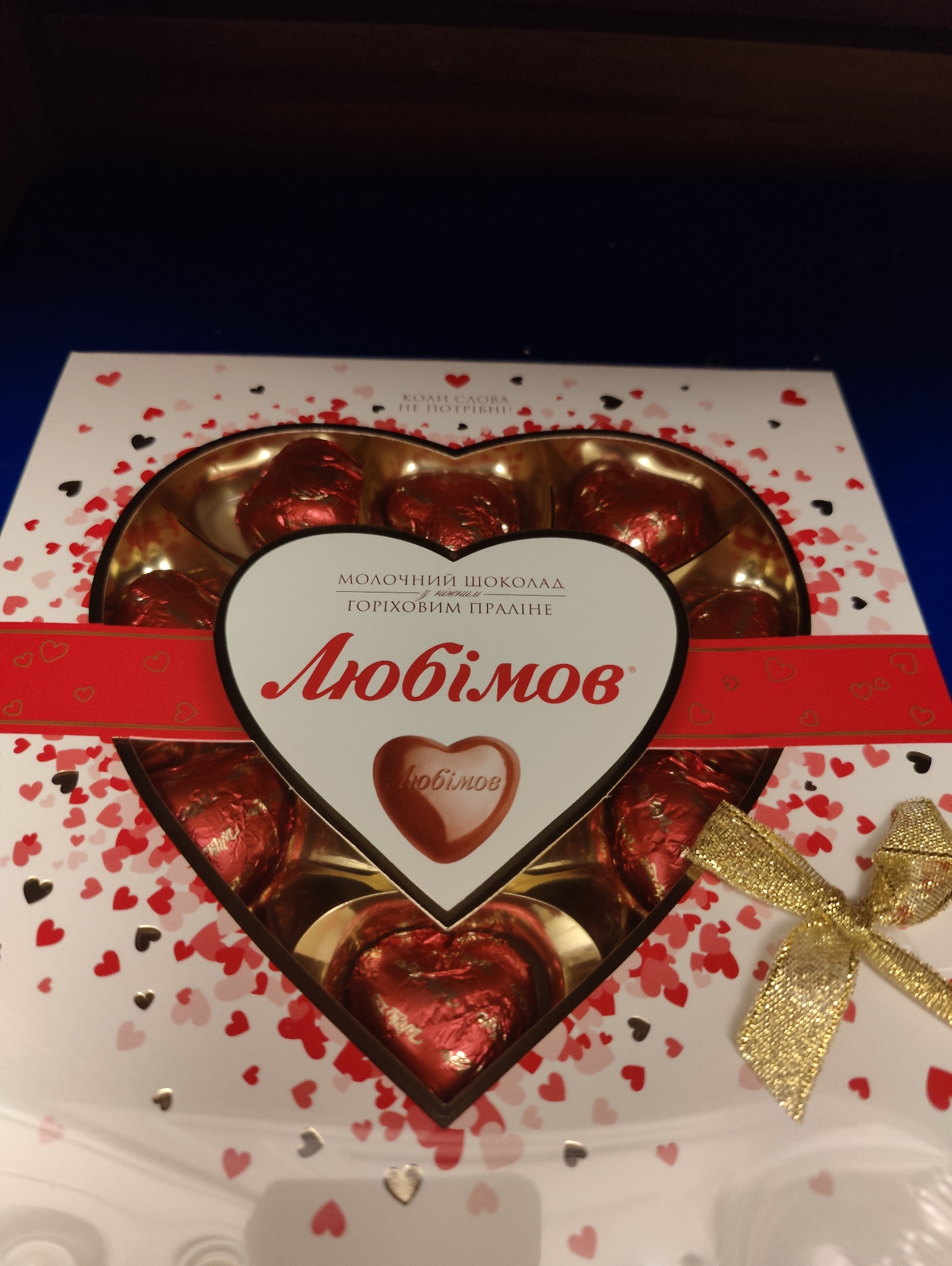 A heart-shaped box of chocolates is displayed on a dark surface, with a blue surface visible in the background. The box is white with a pattern of red and black hearts. A red ribbon with gold trim wraps around the front of the box, adorned with small heart cutouts. In the center of the box, a larger white heart-shaped cutout reveals an arrangement of heart-shaped chocolates. The chocolates are wrapped in shiny red foil. Inside the white heart cutout, text reads "МОЛОЧНИЙ ШОКОЛАД" (milk chocolate) and "ГОРІХОВИМ ПРАЛІНЕ" (nut praline). Below this, the brand name "Любов" is prominently displayed in large red letters, with a smaller heart-shaped chocolate also bearing the name "Любов". A gold bow is tied on the right side of the box.