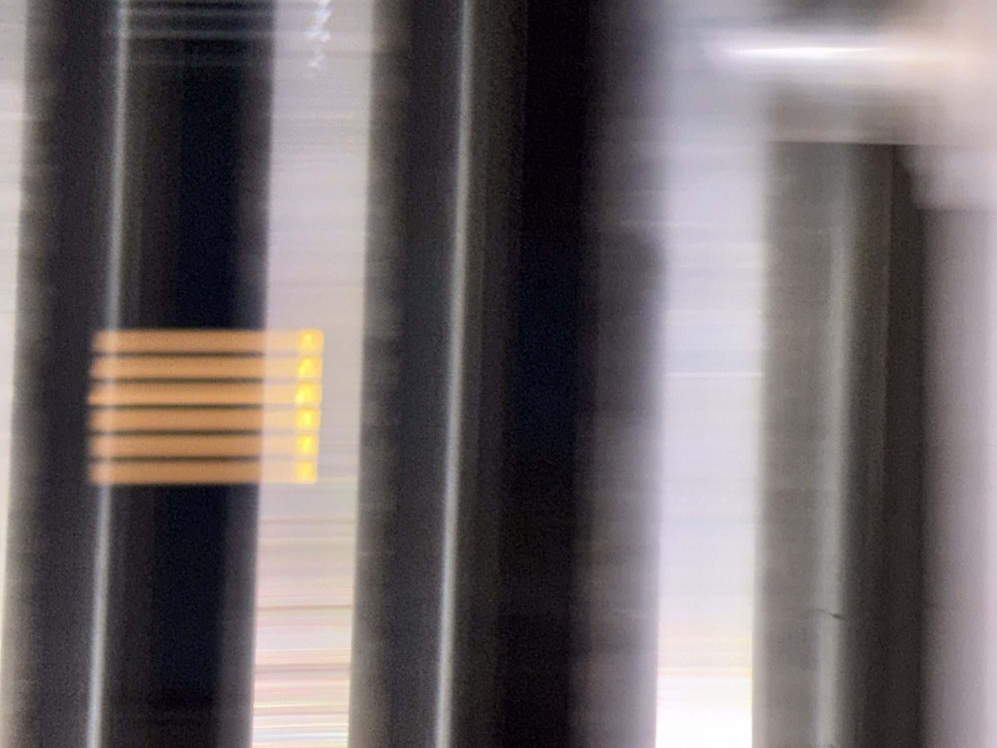The image displays a close-up, slightly blurred view of what appears to be a series of horizontal, dark gray blinds. Between two of these blinds, a segment of thin, vertical orange and yellow lights is visible. These lights are arranged in a row, with alternating orange and yellow illumination. The background is out of focus and appears to be a light-colored, possibly metallic surface with vertical streaks of light. The context of Gempol, Indonesia, is not visually represented or inferable from the image content itself.