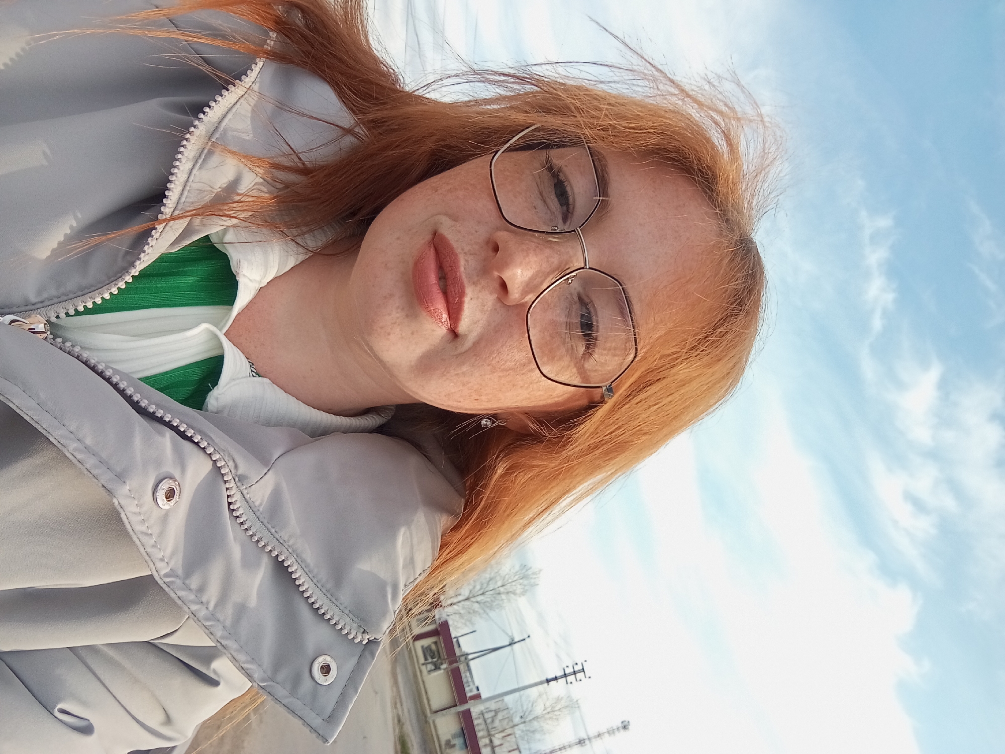 The image shows a woman with red hair and freckles wearing glasses, a white top with a green collar, and a gray jacket. She is outdoors, and the background reveals a blue sky with light clouds and a street scene. A building with a red roof and white walls is visible in the background. There are utility poles and bare trees lining the street. Based on the image context, the location is likely Yurgamysh, Russia.