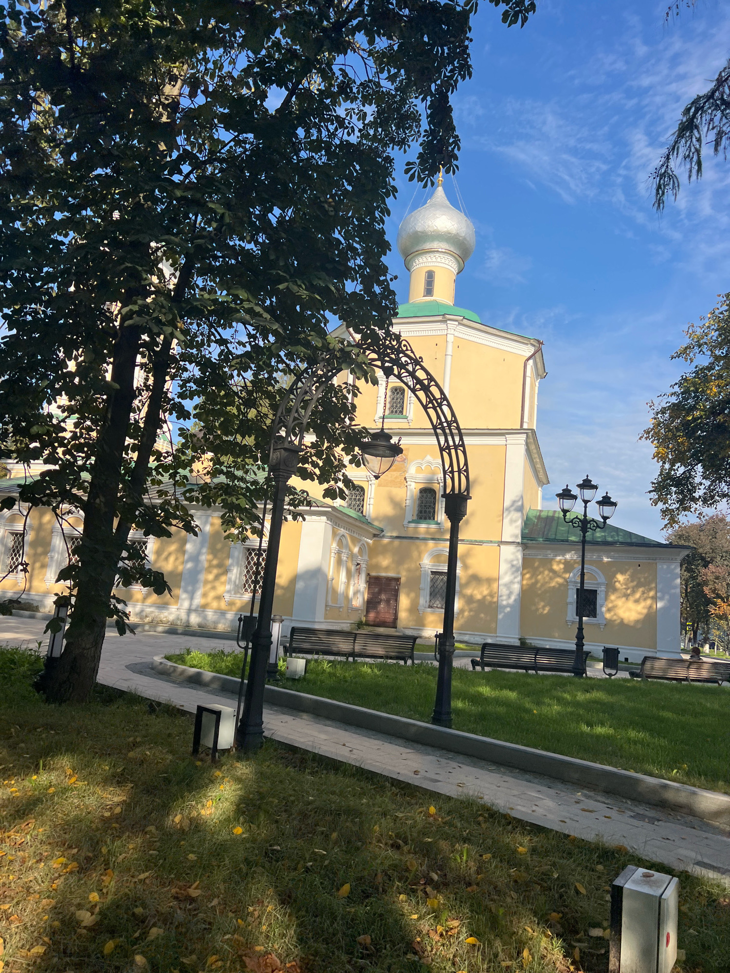 The image shows a sunny day in a park with a yellow building and a silver dome. In the foreground, there is a tree with green leaves and fallen yellow leaves on the grass. A paved walkway leads past the tree. Several dark-colored benches are placed along the walkway. Ornate streetlights are also visible. In the background, the building has a green roof and white trim. The sky is blue with wispy white clouds. The scene appears to be a tranquil outdoor setting, possibly a historical site or a public park, in a city that is not identified. There are no people visible in the immediate foreground, though there is a person seated on a bench in the far right background.