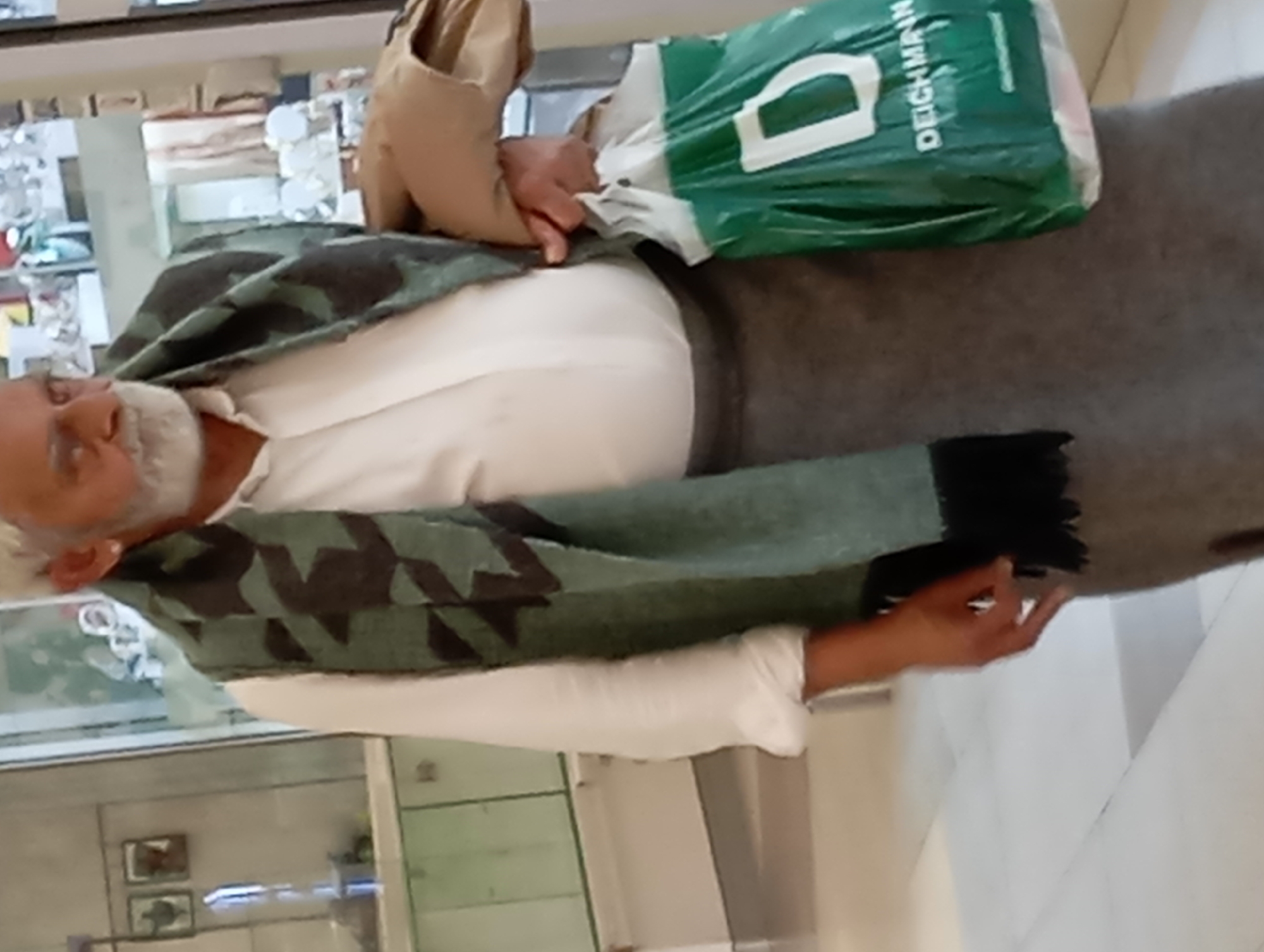 A person, appearing to be a man, stands in an indoor shopping environment, likely a mall. He has a light-colored beard and is wearing a white collared shirt, a dark green scarf with a geometric pattern, and a gray skirt or long coat. He is carrying a green shopping bag from "DEICHMANN" in his right hand. The background shows shop displays and flooring typical of a commercial establishment. The location is identified as the city of Nitra, Slovakia.