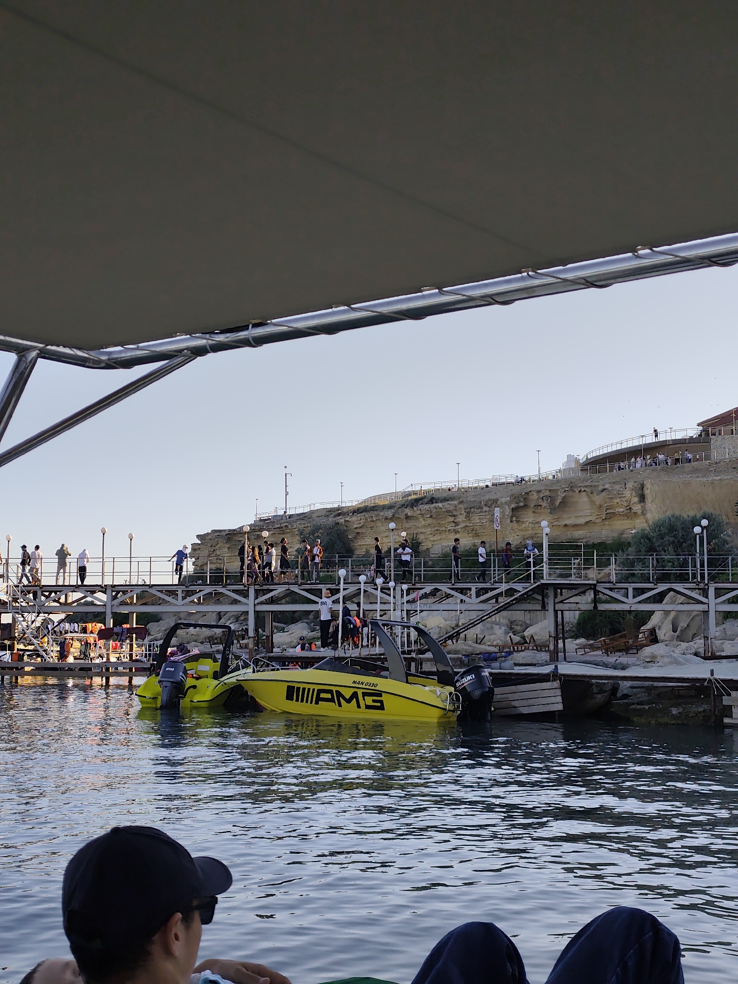 The image depicts a lively scene at a marina, likely in Aktau, Kazakhstan. The foreground shows the legs and back of a person wearing a black baseball cap, seated and looking out towards the water. The focus of the image is the marina, where several boats are docked.
A prominent yellow speedboat with the "AMG" logo and "MAN 0330" text is the most striking object. Another, similar yellow boat is docked next to it. The boats are in the water, with a wooden dock visible behind them.
On the dock and a walkway extending over the water, a group of people are gathered. They appear to be casually strolling and observing the surroundings. The walkway is elevated, providing a view of the marina and the cliffs in the background.
Behind the walkway, a rocky cliff face rises, with a building or structure situated on its summit. The cliff has a weathered, sandy appearance. Scattered vegetation grows on the cliff and around the marina area. The sky is clear and a pale blue, indicating it is likely daytime, possibly late afternoon or early evening given the soft light. The overall atmosphere seems relaxed and leisurely, with people enjoying a day at the waterfront.