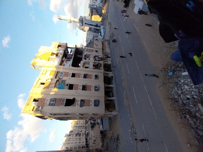 The image presents a severely damaged urban landscape in Khan Yunis, Palestinian Territory, under a partly cloudy blue sky. A multi-story, light-colored residential building, approximately seven to eight floors high, stands centrally, exhibiting extensive structural damage including large holes in its facade, missing windows, exposed interiors, and visible rebar. A blue tarp hangs from an upper-floor opening on the left side of this building. To its right, a white mosque minaret with a pointed cap and a dome at its base is visible amidst rubble and further damaged structures. A two-lane asphalt road with faded white markings occupies the mid-ground. Approximately 10-15 individuals, primarily children and young adults, are scattered across the road and its dirt verges; some are walking, one appears to be on a skateboard, and another is low to the ground, possibly on a scooter or similar object. In the left background, additional multi-story buildings show signs of damage, and open, sandy ground with scattered debris is visible. The foreground consists of a mound of concrete rubble and dirt, suggesting the image was captured from an elevated, possibly damaged, vantage point. Partial view of a dark bag and a leg are visible in the bottom right, confirming the elevated perspective. The scene is illuminated by direct sunlight, casting shadows.