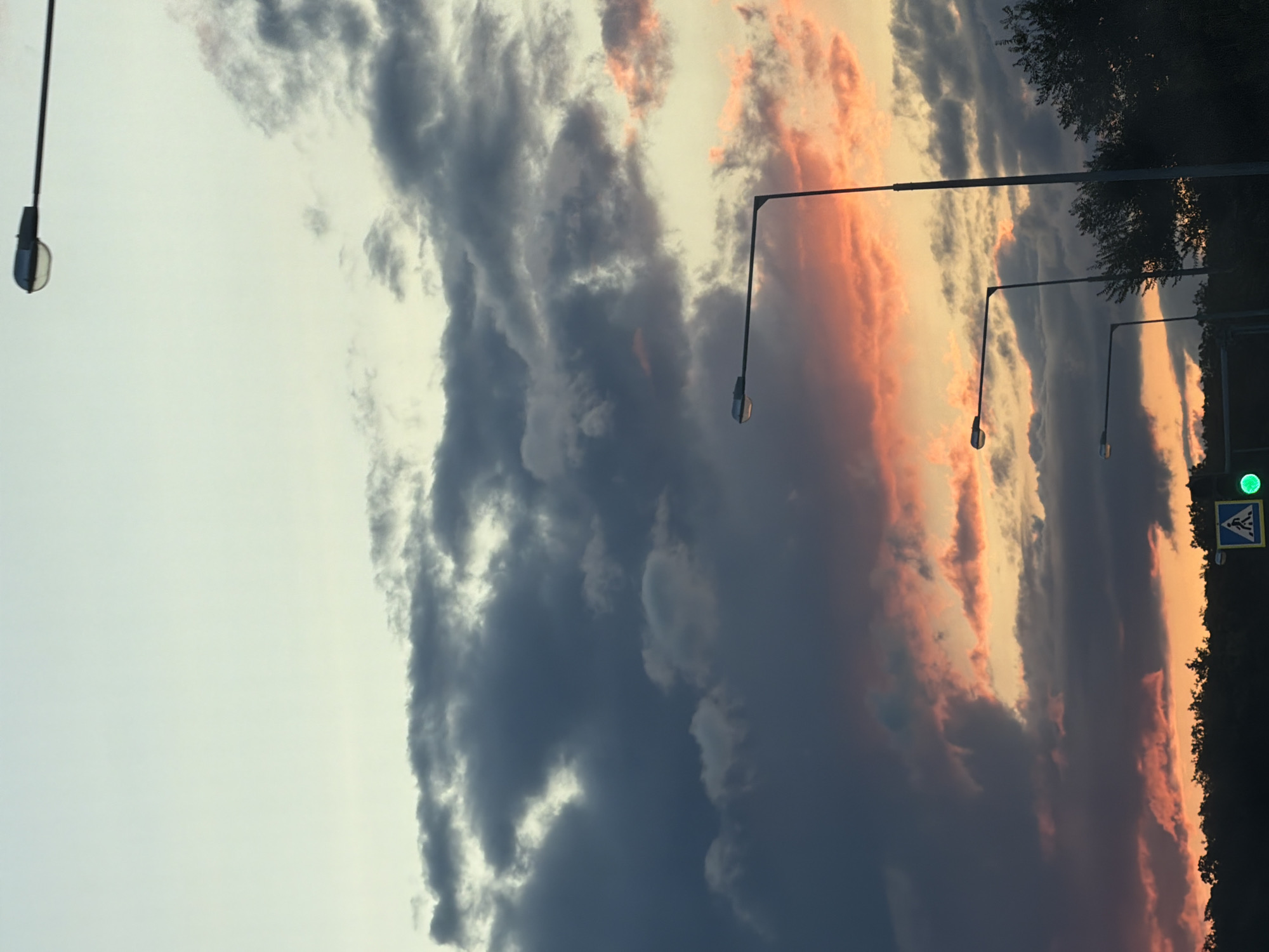 The image shows a sunset over a city, with dramatic clouds lit by the setting sun in shades of orange and pink. Several streetlights are visible, casting long shadows. A pedestrian crossing sign is present on the right side of the image, illuminated by a green light. The dark silhouette of trees lines the horizon. The scene appears to be on a road or street in an urban environment. It is not possible to definitively determine the location as the city of Samara, Russia, without additional context or identifying landmarks.