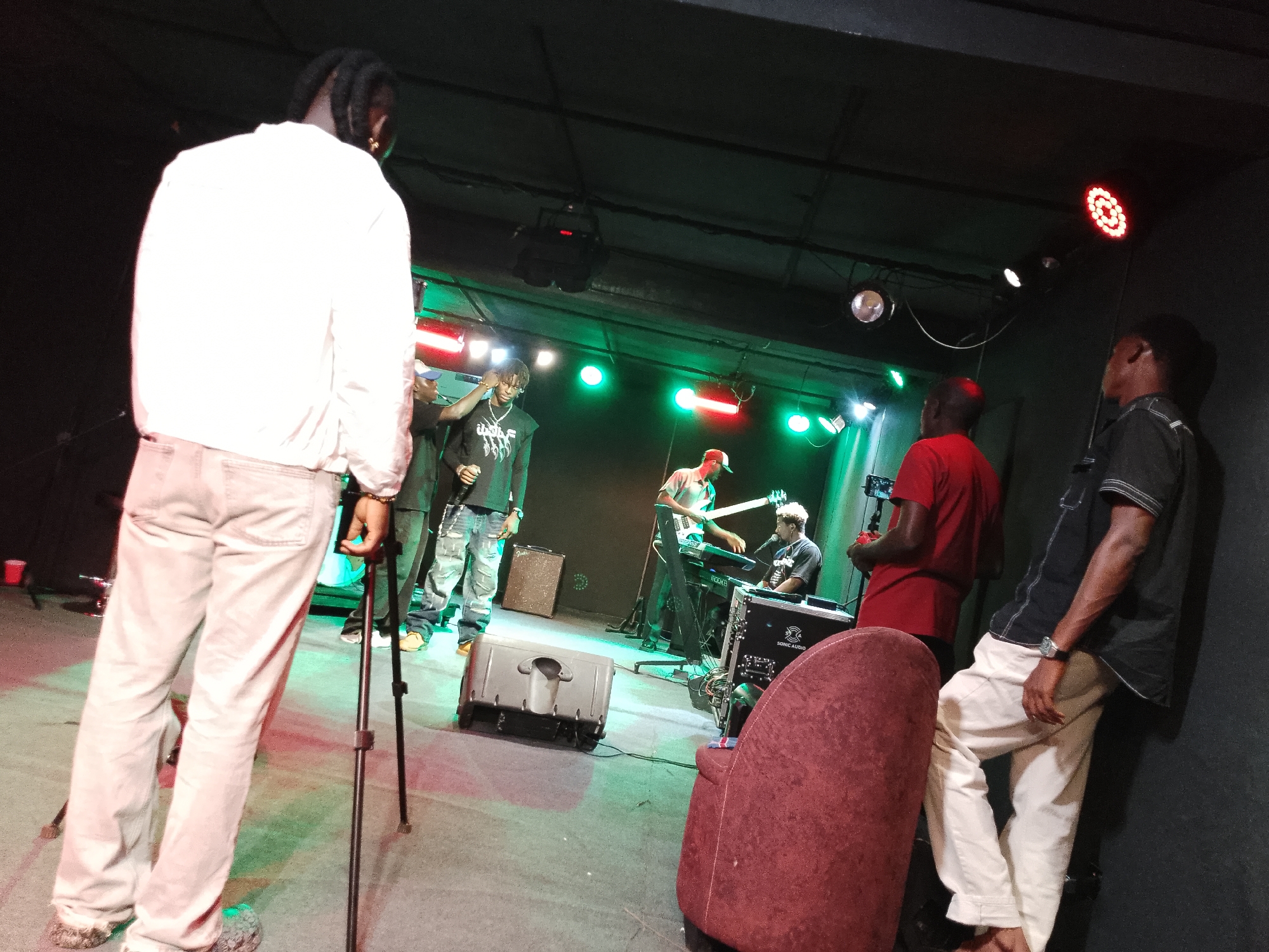 The image captures a dimly lit indoor scene, possibly a music venue, with a band performing on a stage. The primary subject, a person in a white jacket and light-colored pants, stands with their back to the camera, facing the stage. They are positioned on the left side of the frame, with a tripod standing to their right.
On the stage, several individuals are actively engaged in music. A person wearing a black graphic t-shirt and jeans is singing into a microphone, while another individual plays a white electric guitar. To their right, a keyboard player is seated. Stage lighting illuminates the performers with green and red hues, creating a vibrant atmosphere.
Various musical equipment is visible, including speakers, amplifiers, and electronic devices. A plush red chair is situated in the foreground on the right side of the image, partially obscuring the view of one of the band members. The background consists of a dark, textured wall and ceiling, punctuated by spotlights. The overall impression is that of a live music performance in progress, with an audience member observing from the side.