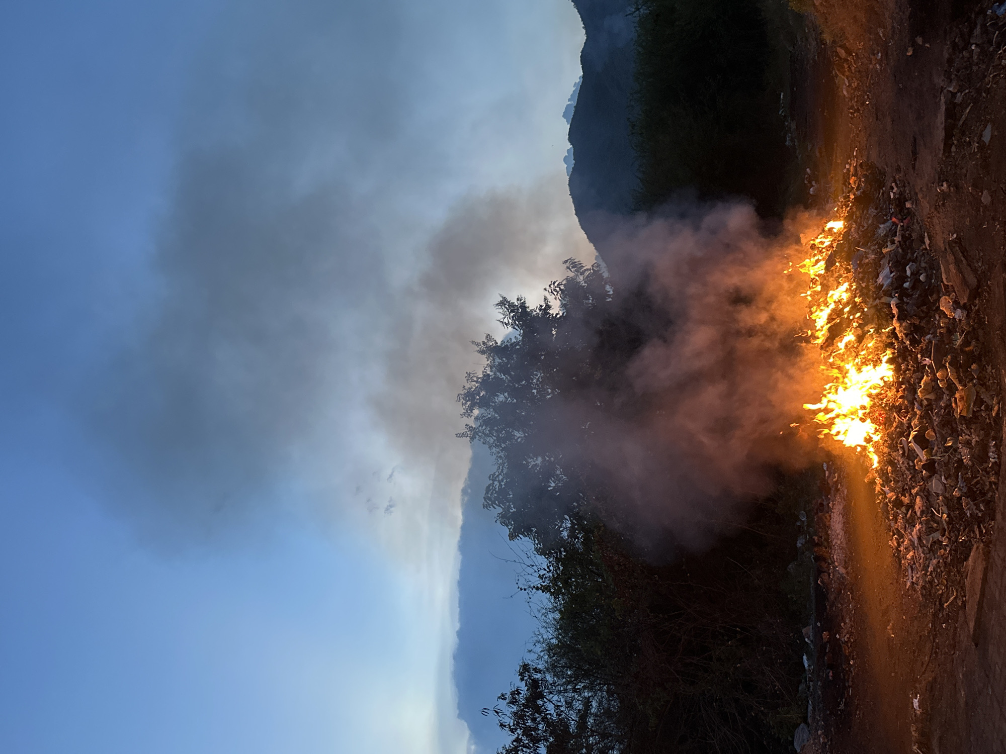 The image shows a fire burning beside a road in Bac Nha Trang Ward, Vietnam. The flames are visible on the right side of the image, with smoke rising and partially obscuring trees in the background. The sky is visible on the left side, with a transition from blue to darker clouds. A mountain is visible in the distance behind the trees. The foreground consists of a dirt road.