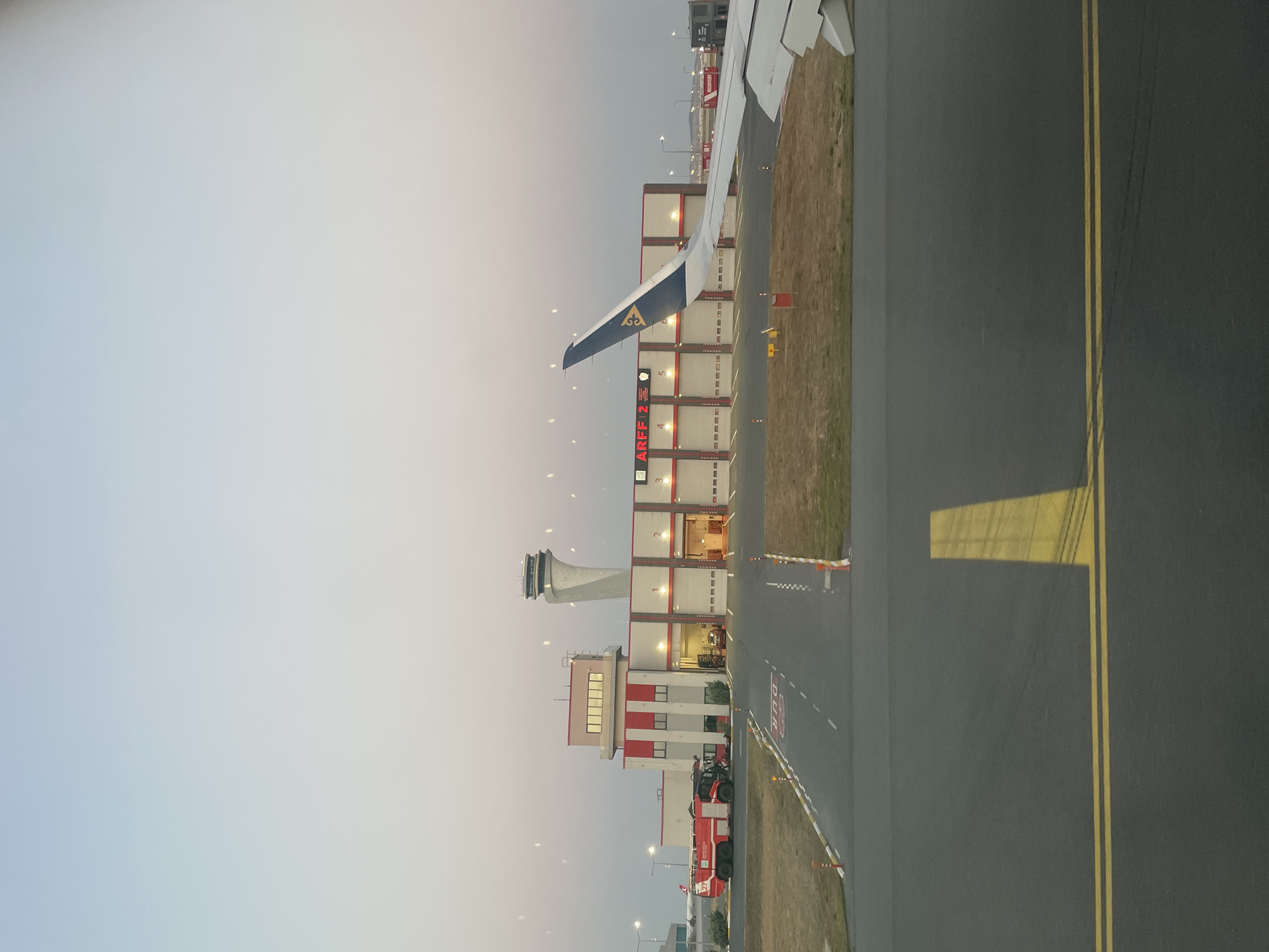 An airport building with the text "ARFF 2" is visible, along with a control tower in the background and an airplane wing in the foreground. The building is red and white, with several garage doors. A red fire truck is parked in front of the building. The scene appears to be an airport tarmac, possibly at sunrise or sunset, given the soft lighting. The image does not provide specific details that would confirm the location in Beykoz, Turkey.