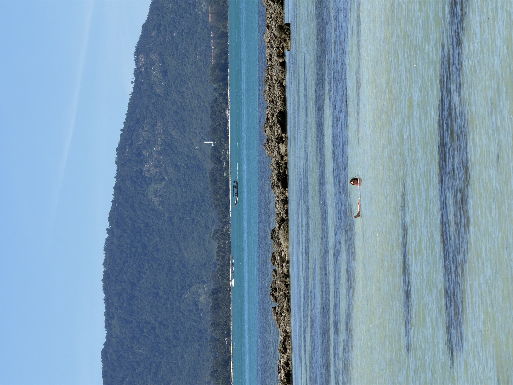 The image depicts a serene coastal scene in Baan Thong Krut, Thailand. The composition is dominated by a lush, verdant mountain slope that recedes into the distance on the left side of the frame. The mountain is densely covered with trees, suggesting a tropical environment.
The ocean occupies the central and right portions of the image. The water is a vibrant turquoise, with clear visibility revealing streaks of lighter and darker blue, likely due to variations in depth and the play of sunlight on the seabed. A rocky outcrop juts out from the sea, creating a textured contrast with the smooth water.
In the foreground, on the right side of the image, a person is partially submerged in the water, appearing to be swimming or wading. They are looking towards the left, with only their head and upper body visible. The water is calm, with gentle ripples reflecting the sunlight.
Further out in the ocean, several boats are visible. One is a larger vessel with a dark hull, while others appear to be smaller, possibly traditional Thai longtail boats. The presence of these boats suggests a location frequented by tourists or locals engaged in water activities.
The sky is a clear, bright blue, indicating a sunny day with no clouds. The time of day appears to be midday or early afternoon, given the intensity of the sunlight. The overall atmosphere is peaceful and idyllic, capturing the essence of a tropical beach holiday. There is no visible text in the image.