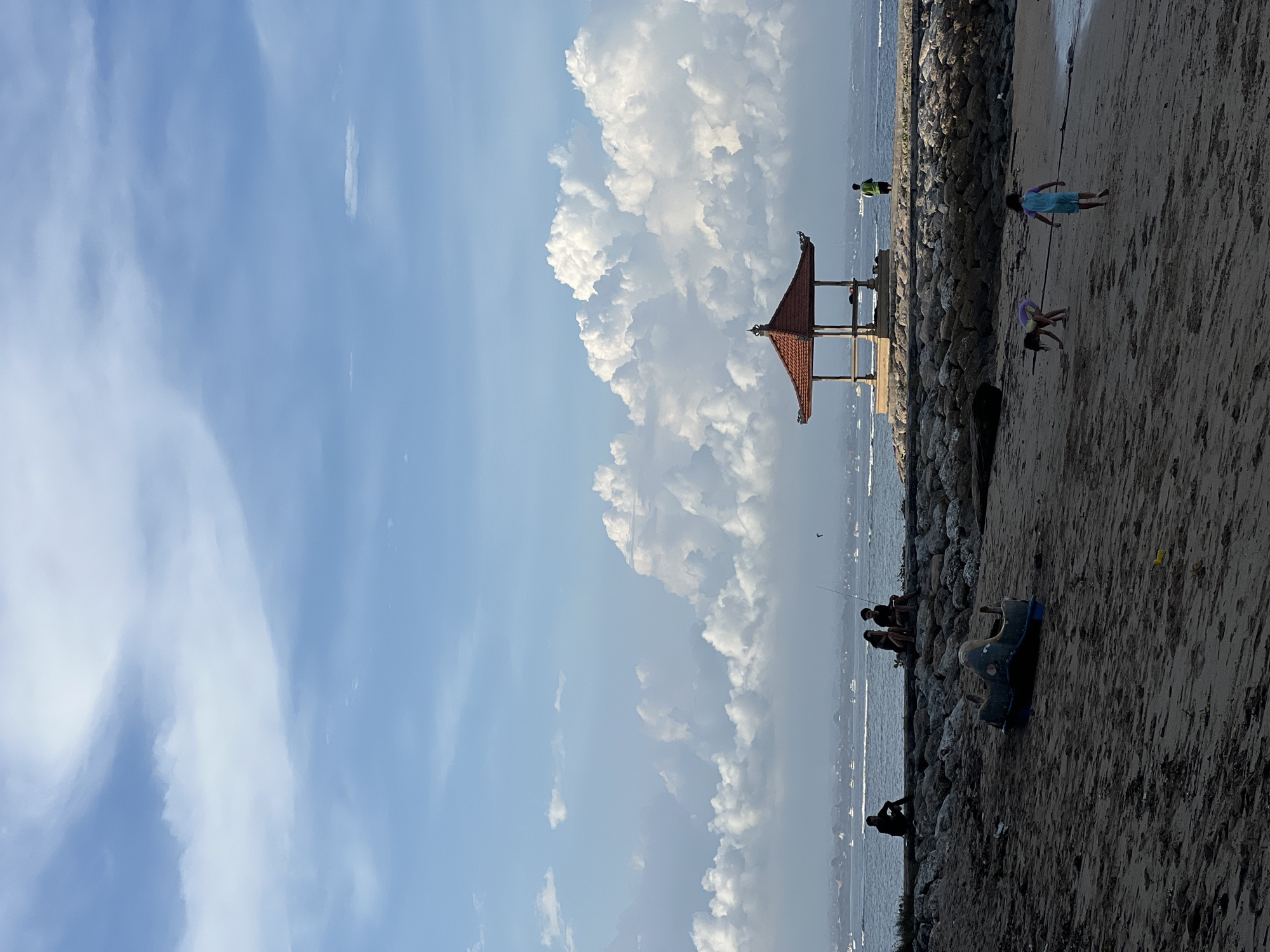 The image shows a beach in Denpasar, Indonesia. The sky is partly cloudy. In the background is a gazebo. On the beach are children playing, people fishing, and discarded toys. The picture is taken at an angle.