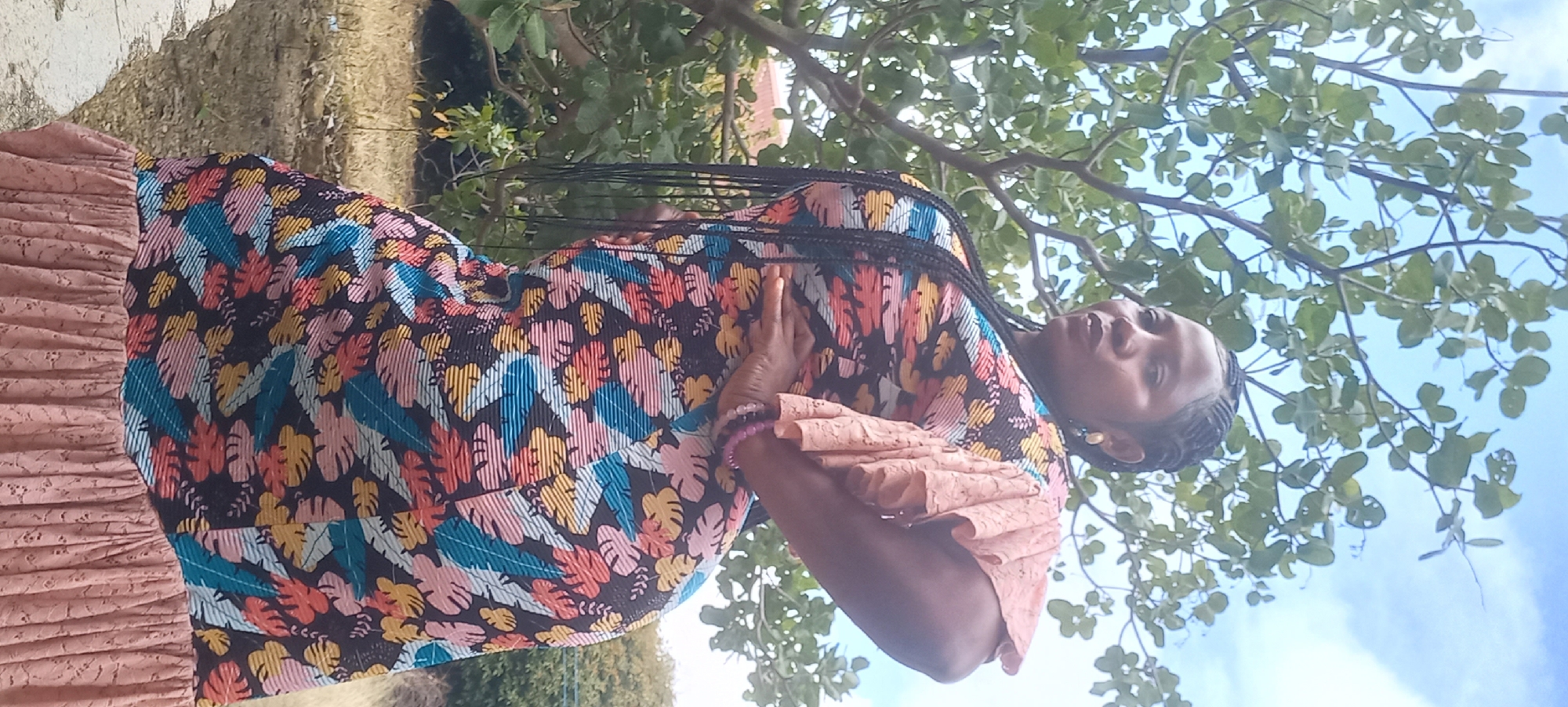 A Black woman with braided hair stands outdoors in front of a tree with green leaves and a pale blue sky with white clouds. She wears a dress with a colorful floral pattern and ruffled, peach-colored sleeves. Her left hand rests on her hip, and her right arm is bent with her hand on her stomach. She is wearing earrings and a bracelet. The background shows dry grass and foliage, with hints of buildings in the distance. The scene appears to be in Jos, Nigeria.