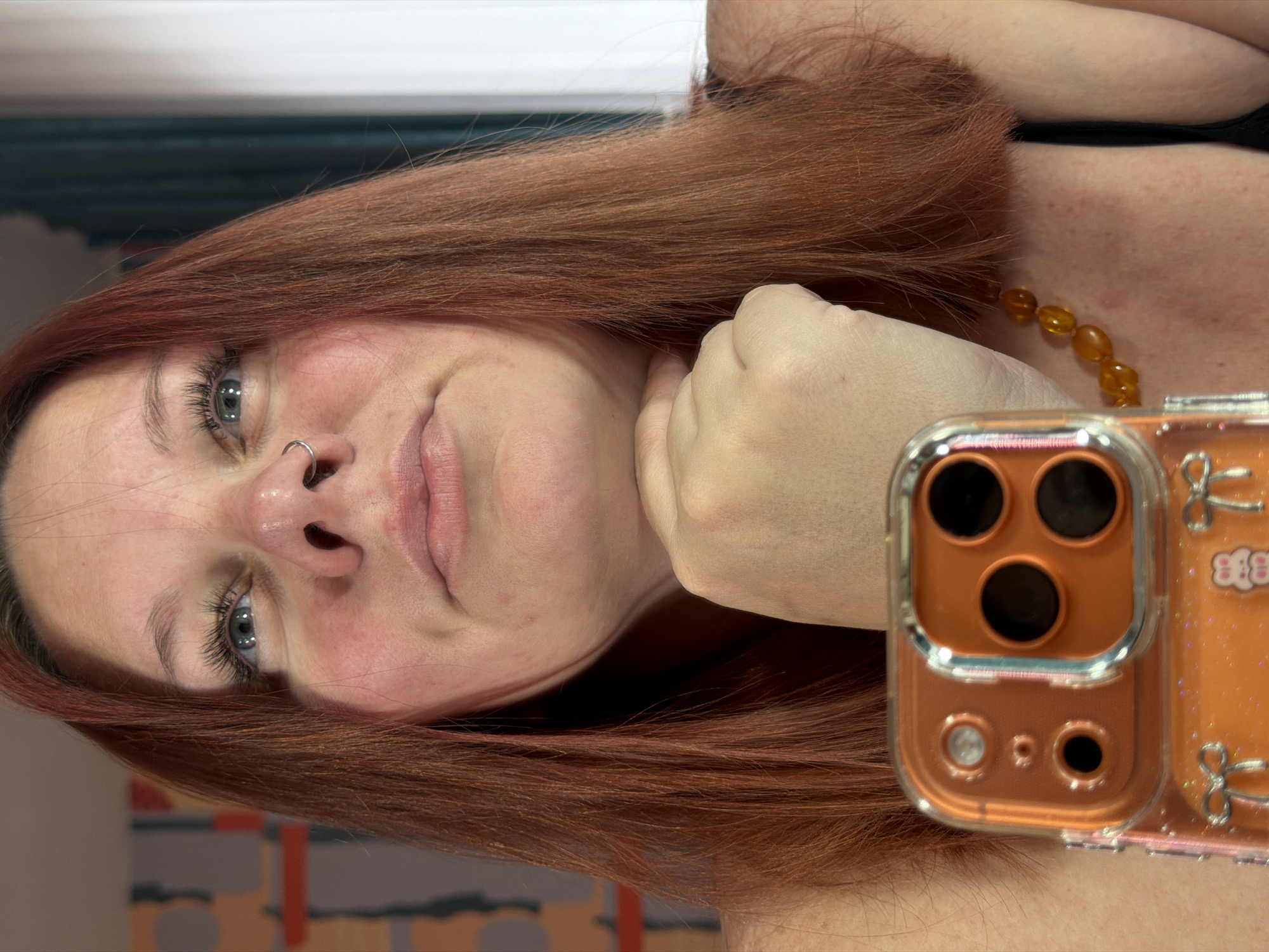 A selfie taken by a person with shoulder-length reddish-brown hair. The person has light skin, blue eyes, and a nose ring. They are wearing an amber beaded necklace and appear to be wearing a black top. The person is holding an orange phone case with a butterfly design up to their face. The background is blurred, but there appears to be a window with blue curtains and a wall with a colorful abstract design. No evidence suggests the image was taken in the city of Chaikovsky, Russia.