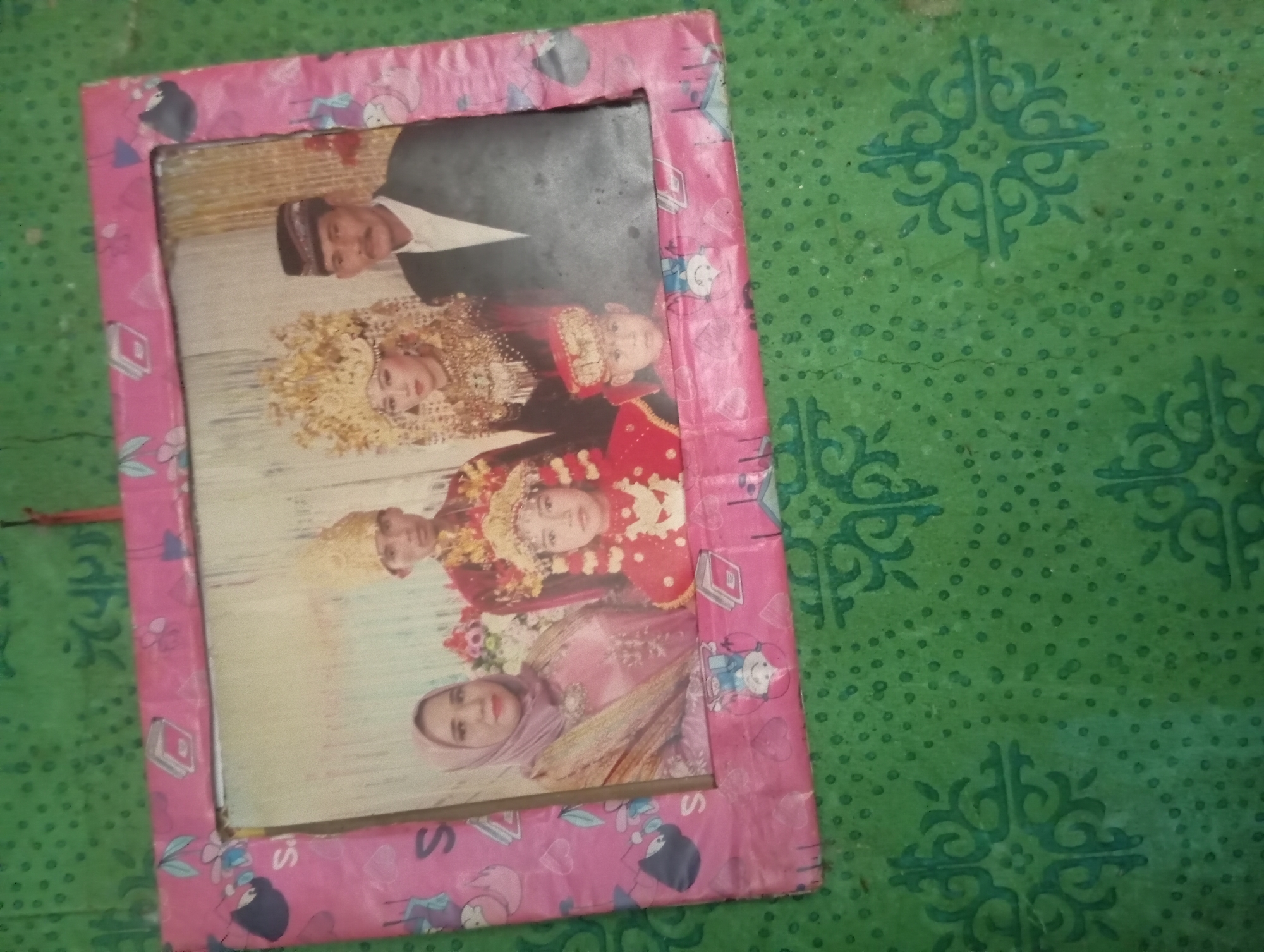 The image shows a framed photograph of a family, possibly during a wedding ceremony, displayed against a green patterned background. The photograph features at least five individuals. In the center, a young woman wears an elaborate golden headdress and ornate jewelry, suggesting she is the bride. To her right stands a man in a dark jacket and a traditional cap, likely the groom. To the bride's left, a woman in a light pink hijab and a matching dress is visible, possibly a mother or relative. In front of the bride and groom, a young boy wears a red cap and a red outfit. The photograph is enclosed in a pink frame decorated with cartoon-like images of birds and possibly other figures. The green background appears to be a textile or a wall covering with a repeating ornamental pattern of teal swirls and dots. The overall impression is of a personal memento displayed in a domestic setting. The image's quality suggests it may be an older photograph. The location of Rejosari, Indonesia, is mentioned, but there are no visual cues within the image itself to confirm this specific location.
