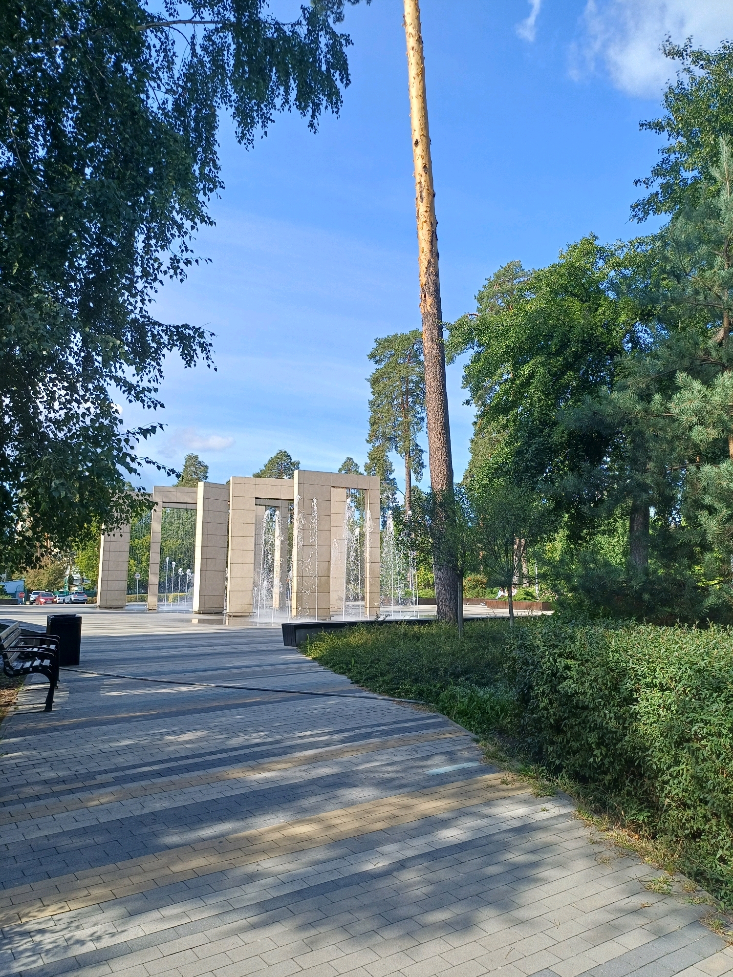 The image depicts a public park area in Kyiv, Ukraine. In the foreground, a paved walkway with a pattern of light and dark gray rectangular stones is visible, casting long shadows from overhead trees. A dark park bench and a trash receptacle are situated to the left of the walkway. Beyond the walkway, a modern fountain installation with multiple tall, rectangular pillars is featured, with water spraying upwards from between them. Lush green bushes and trees form the background, with a clear blue sky above, dotted with a few white clouds. A tall, slender pine tree stands prominently on the right side of the frame. The overall scene suggests a well-maintained urban recreational space on a sunny day.