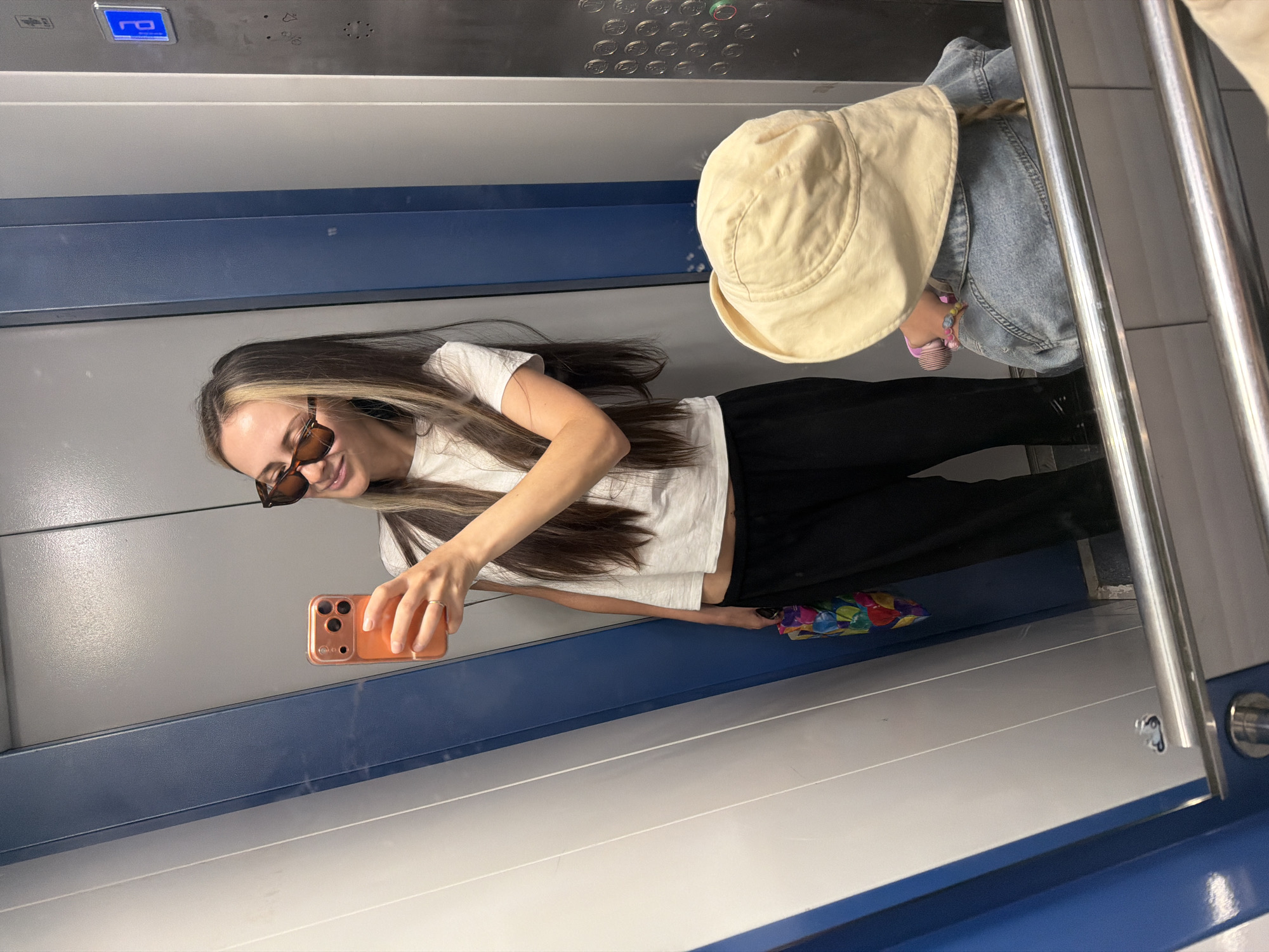The image appears to be a selfie taken in an elevator. A woman with long brown hair is holding an orange smartphone and smiling at the camera. She is wearing a white t-shirt, black pants, and sunglasses. In the background, there is a child in a denim jacket and a light yellow sun hat. The elevator has blue and white panels and a steel handrail. A colorful umbrella is visible on the floor. The elevator buttons and other mechanical components are visible in the background. The location is not possible to ascertain from the image.