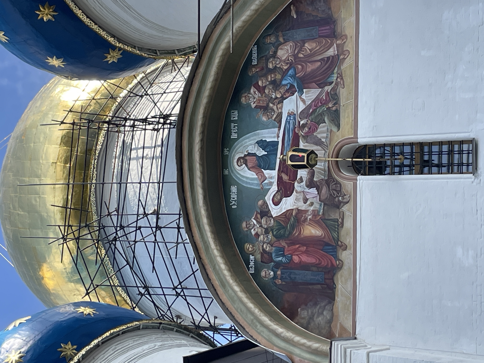 This image captures a detailed fresco adorning the exterior of a church in Sergiyev Posad, Russia. The fresco, set within an arched frame, depicts a religious scene with multiple figures. In the center, Jesus Christ is shown holding the infant Mary, while a deceased figure, likely the Virgin Mary, lies on a bier. Several individuals, appearing to be disciples or mourners, surround the bier, some with expressions of grief. An angelic figure kneels at the foot of the bier, and another figure is seen reaching towards the deceased. Above the scene, Greek inscriptions are visible. The fresco is painted on a white brick facade. In the upper portion of the image, the golden dome of the church, partially covered by scaffolding, is visible alongside a blue dome adorned with golden stars. A tall, narrow window with a metal grate is set into the white facade below the fresco.