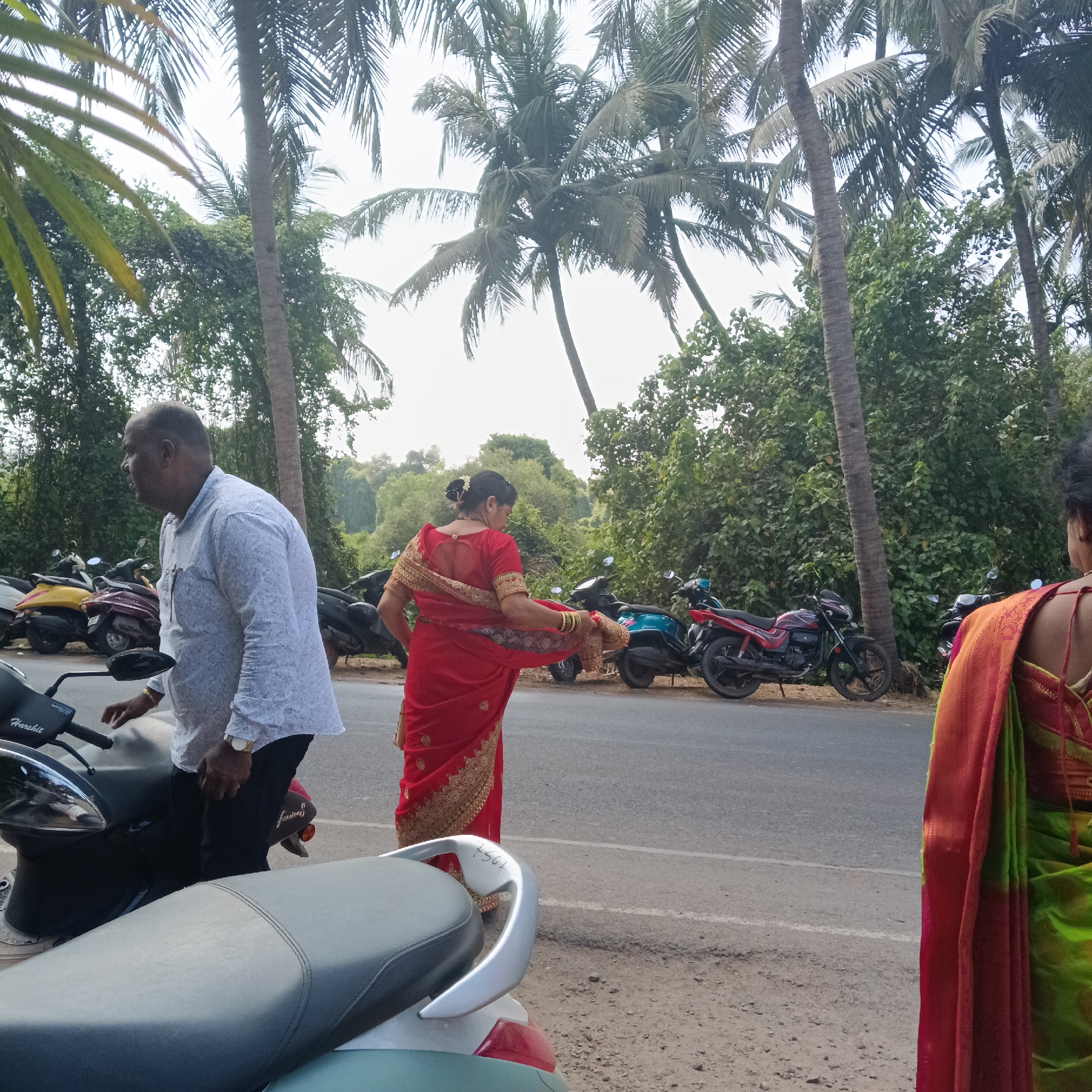 The photograph captures a scene in Mapusa, India, near a street lined with parked motorcycles. A man in a light-colored shirt and dark pants stands next to a scooter, facing left. A woman wearing a red sari stands near the scooters, facing away with her sari draped over her arm. A second woman, partially visible on the right, wears a green sari. Tall palm trees and dense green foliage fill the background. The sky is overcast.