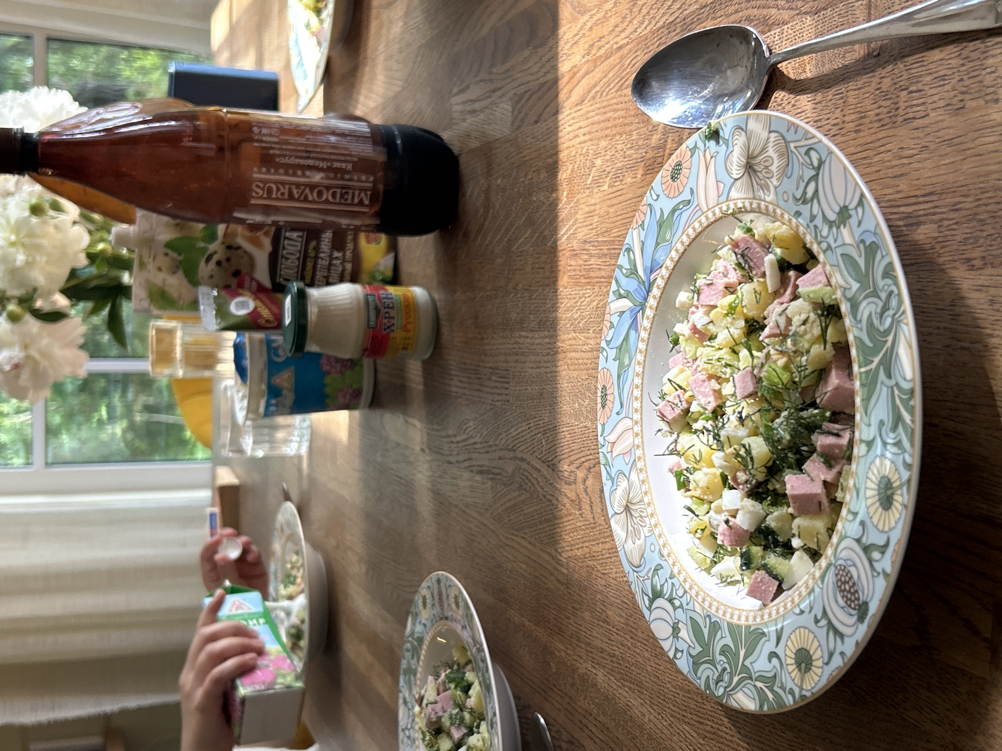 A close-up shot of a wooden table set for a meal, with a focus on two plates of salad and a large brown bottle of beverage. A person's hands are visible in the upper left corner, pouring liquid from a pink and green carton into one of the salad plates. The salad appears to be a mixture of diced potatoes, eggs, ham, and herbs, garnished with dill. The plates have a decorative floral pattern with blue and green accents. On the table, there are also several smaller containers, a glass of water, and a large metal spoon to the right of the main salad plate. In the background, a bouquet of white flowers is visible on the table, and through a window, greenery and a partially obscured outdoor scene suggest a natural setting. The overall lighting indicates a bright, possibly sunny, indoor environment. This scene is likely occurring in a home in Shepelyovo, Russia, a small village within the Leningrad Oblast known for its natural surroundings and historical sites.