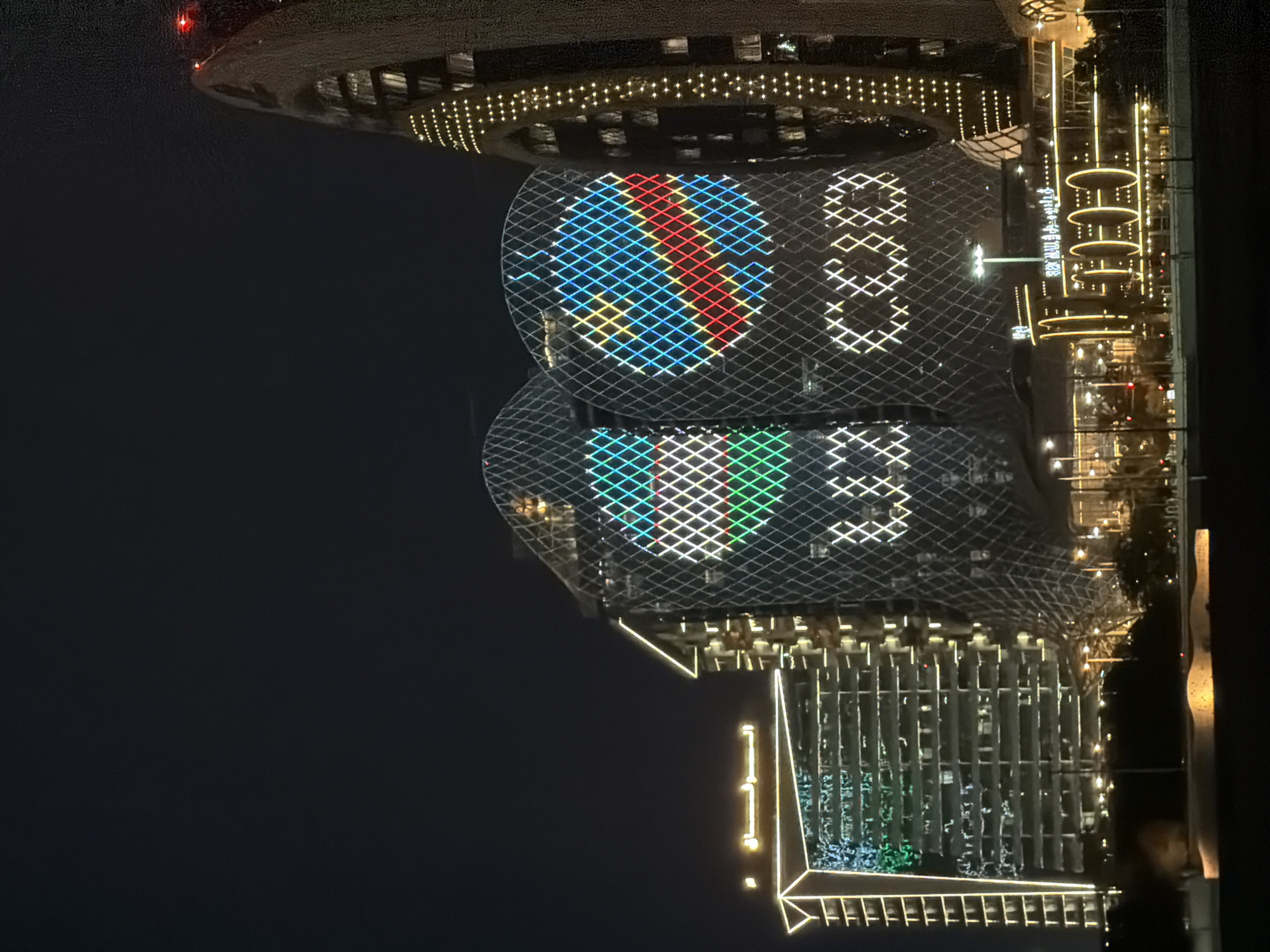 The image depicts a nighttime scene of a city skyline with several modern buildings. The most prominent structures are two cylindrical towers with a diamond-shaped lattice exterior. These towers are illuminated with colored lights, forming patterns that resemble flags and logos. The left tower displays a flag with green, red, and blue horizontal stripes, and the right tower shows a circular logo composed of blue, yellow, red, and green sections. Below these towers, a lower building is visible with Arabic text and yellow circular lights. Further to the right, another tall, slender building is also illuminated with numerous small, yellow lights, creating a vertical pattern. The overall environment is dark, indicating nighttime, with some faint lights visible in the distance. The scene appears to be a vibrant urban landscape at night, likely showcasing a significant architectural or event display.